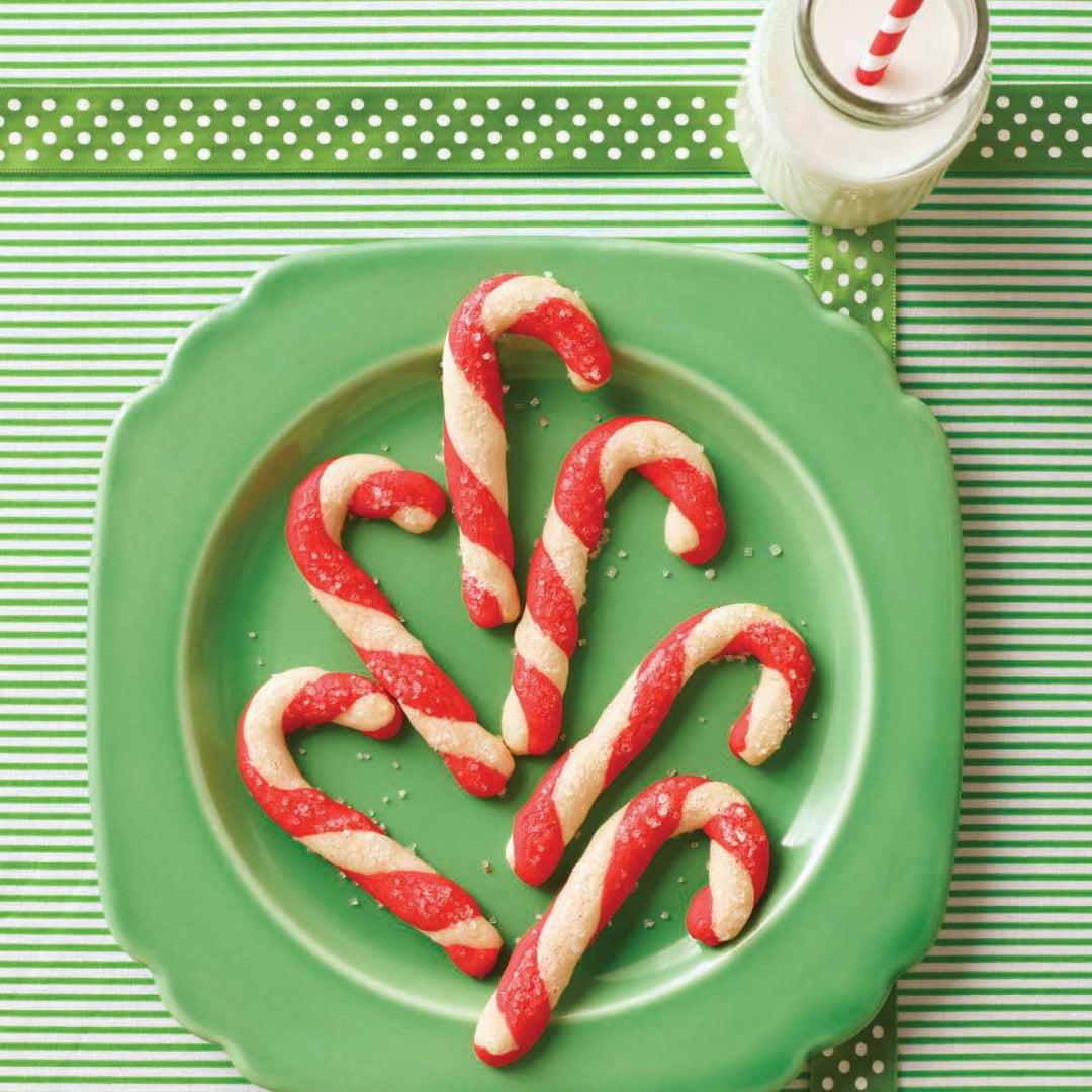 Candy cane-shaped cookies on a green plate with a glass of milk on a striped green and white background.
