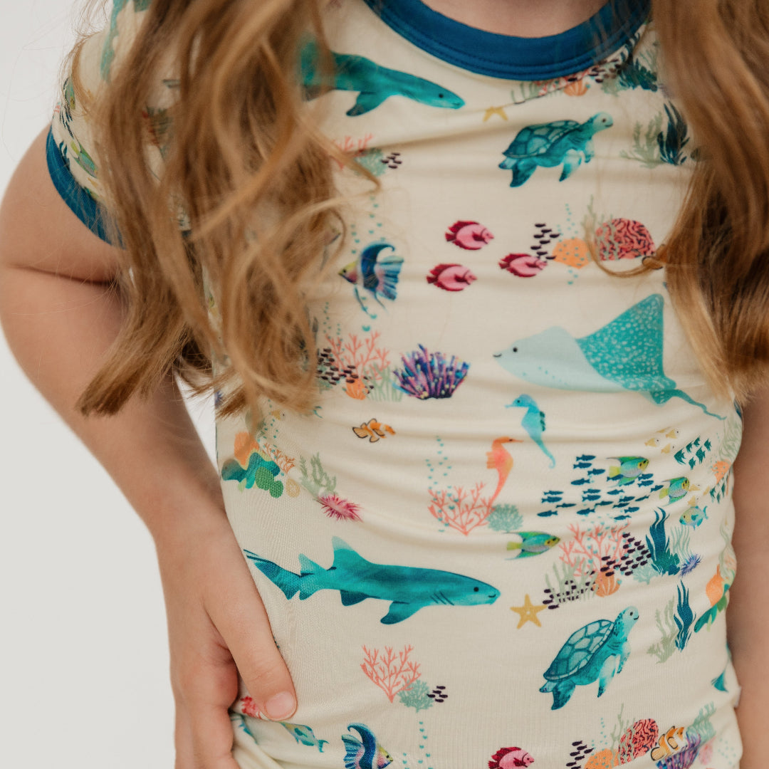Close up of toddler girl wearing Emerson and Friends Coral Reef Bamboo Kids Pajama Set featuring a soft, breathable short sleeve top and matching pants with an ocean-inspired print of sharks, sea turtles, and coral reefs—perfect for cozy nights and sweet dreams.