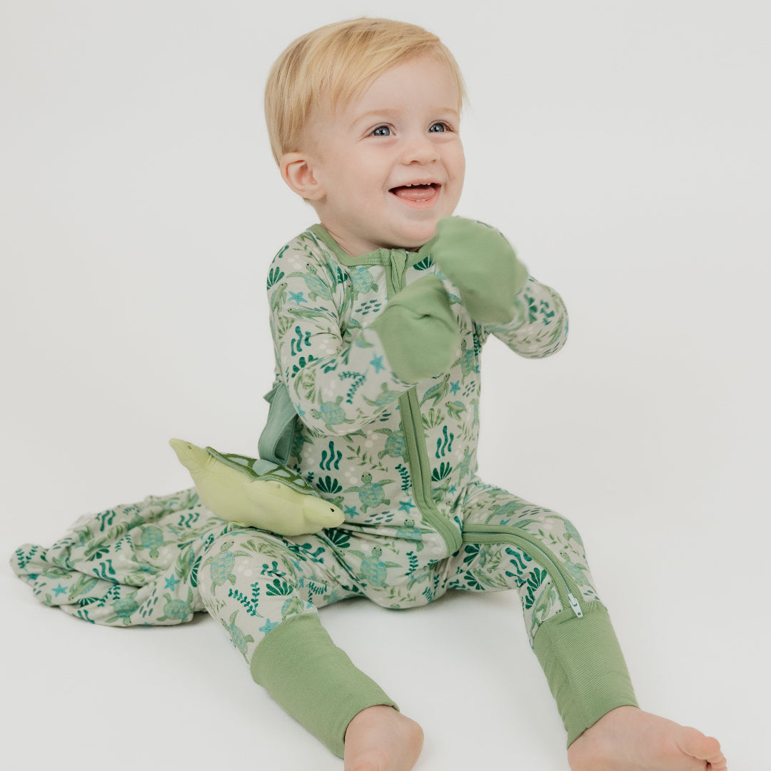 A toddler sitting in a white background with our Save the Sea Turtle lovey, featuring a plush turtle attached to a cozy, ocean-inspired printed blankie. Made from ultra-soft bamboo, this comforting lovey is perfect for snuggles and soothing moments.