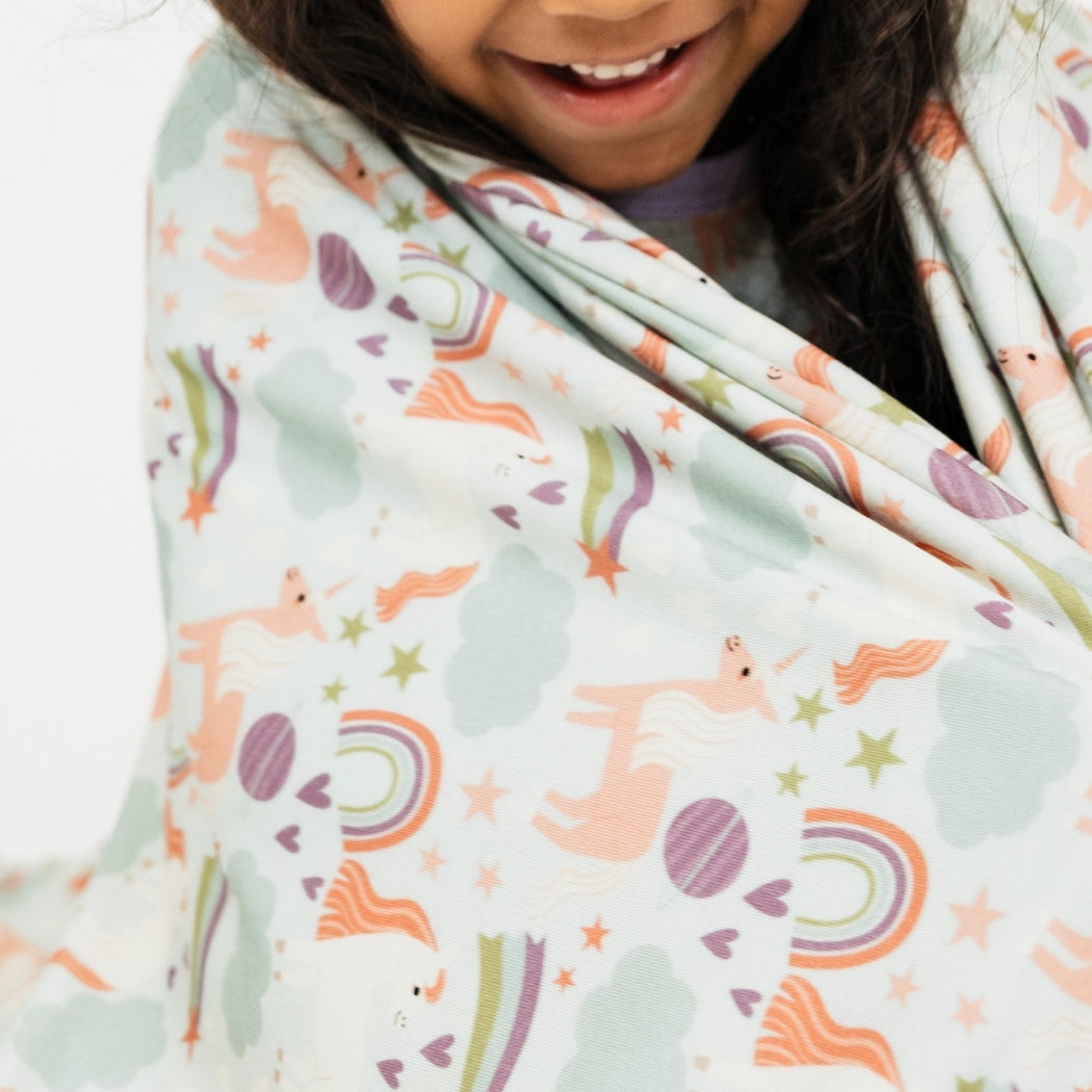 Little girl wrapped up in our Magical Unicorn blanket. The print includes unicorns, rainbows, clouds, planets, stars, and hearts. The colors are purple, pink, and blu, with hints of green.