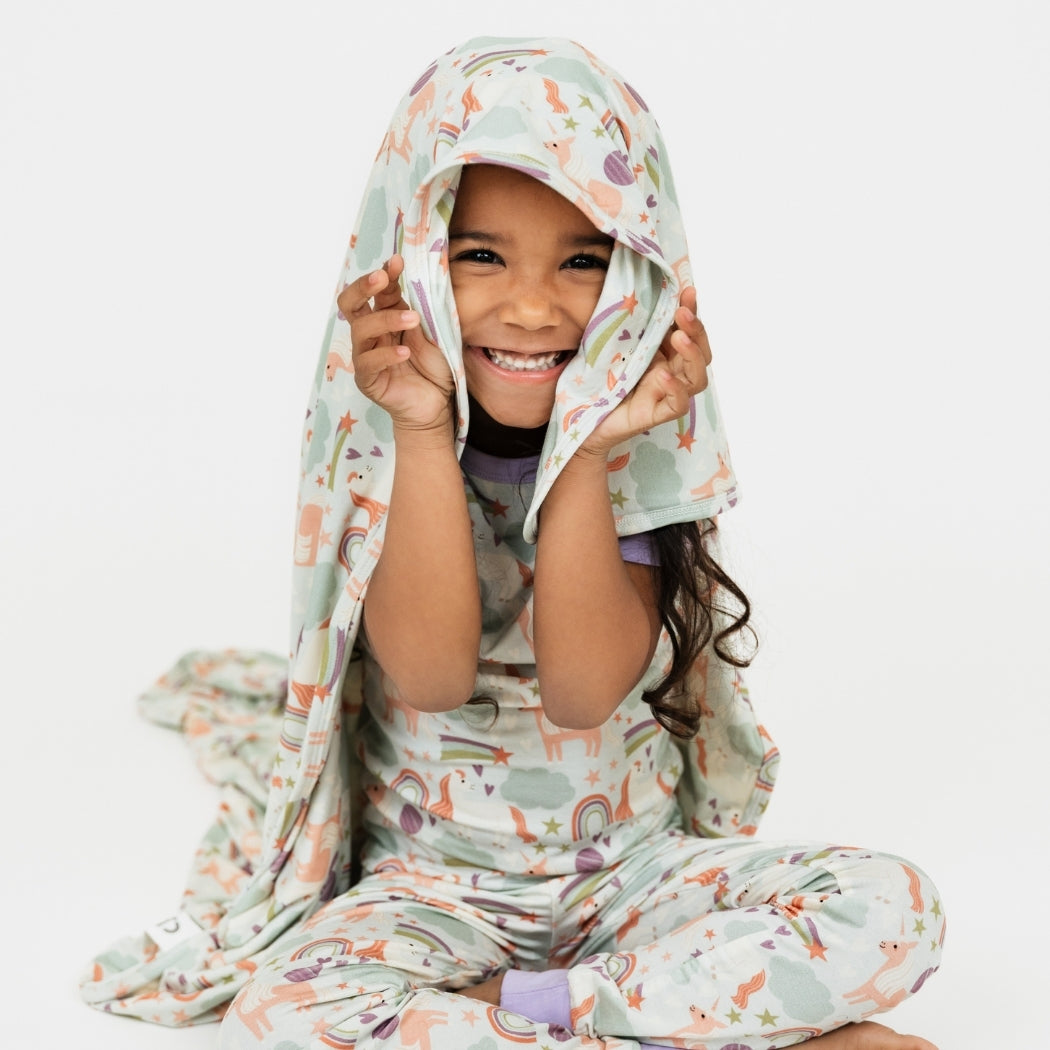 Little girl wearing her Magical Unicorn pajama set, with our Magical Unicorn blanket over her head. The print includes unicorns, rainbows, clouds, planets, stars, and hearts. The colors are purple, pink, and blu, with hints of green.