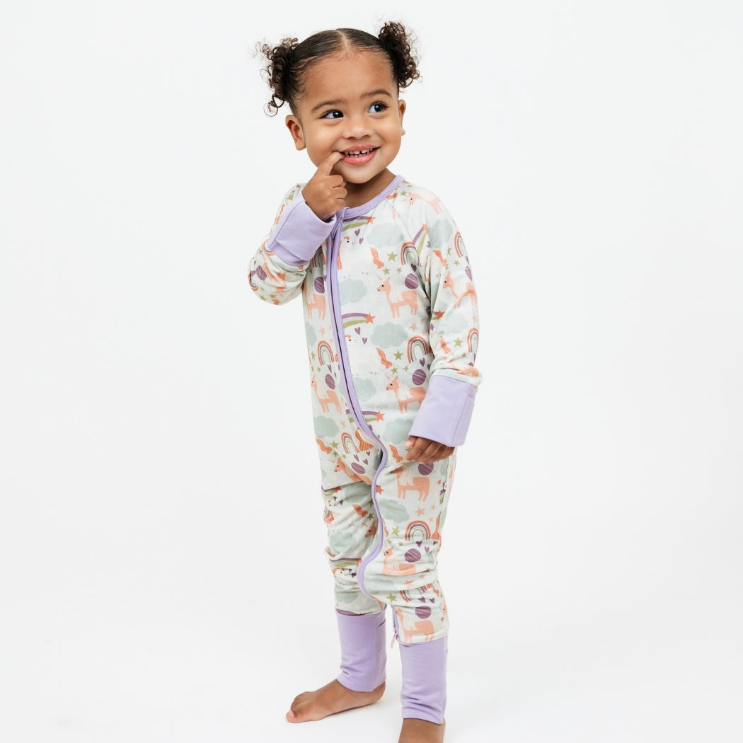 Little girl smiling with her pointer finger in mouth. She is standing and wearing our Magical Unicorn printed bamboo convertible. The print includes unicorns, rainbows, clouds, planets, stars, and hearts. The colors are purple, pink, and blu, with hints of green.