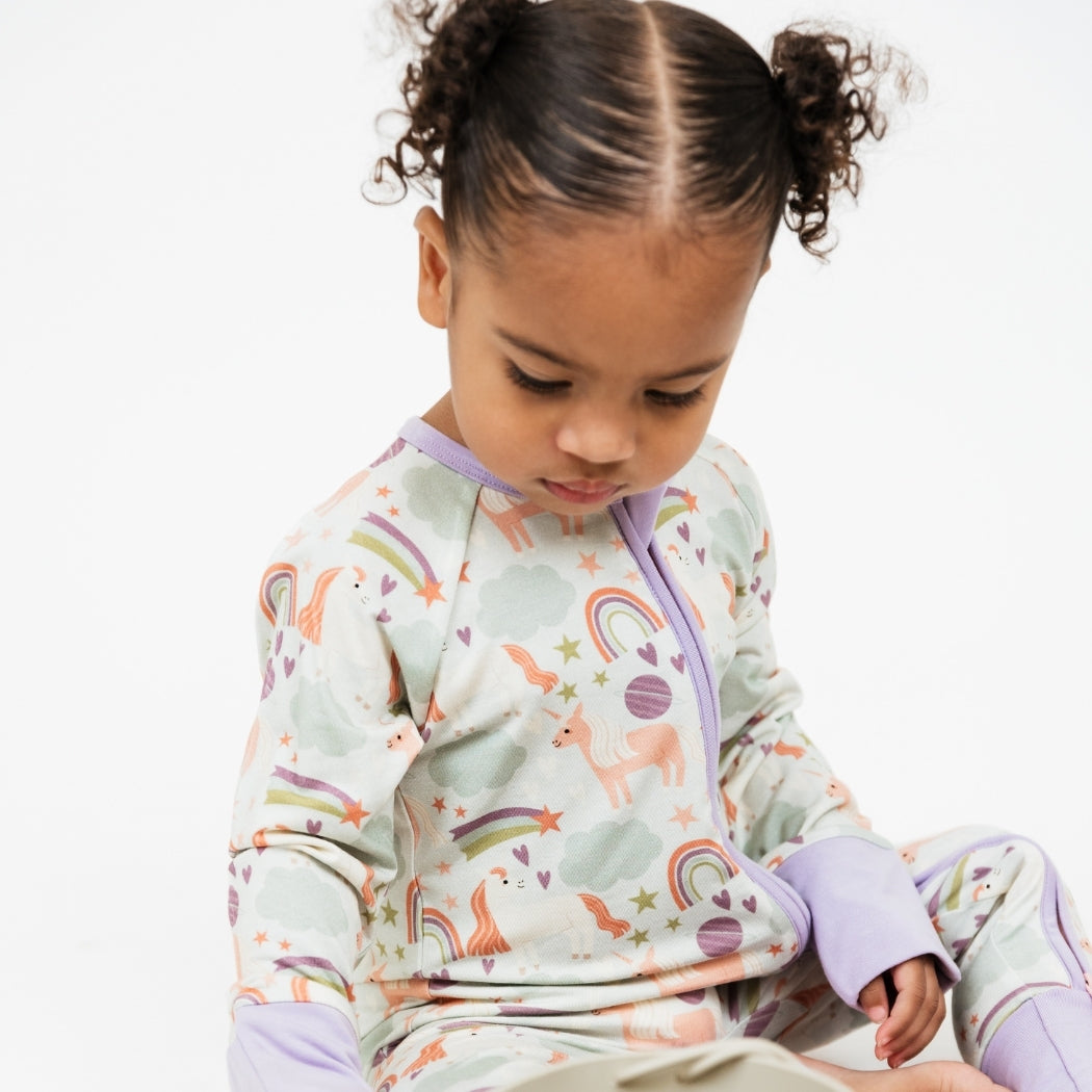 Little girl looking down while wearing our Magical Unicorn bamboo pajama. The print includes unicorns, rainbows, clouds, planets, stars, and hearts. The colors are purple, pink, and blu, with hints of green.