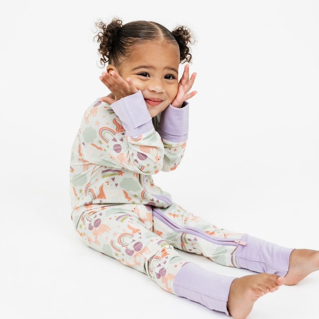 Little girl sitting down in our Magical Unicorn bamboo convertible pajama with her hands on her face smiling. The print includes unicorns, rainbows, clouds, planets, stars, and hearts. The colors are purple, pink, and blu, with hints of green.