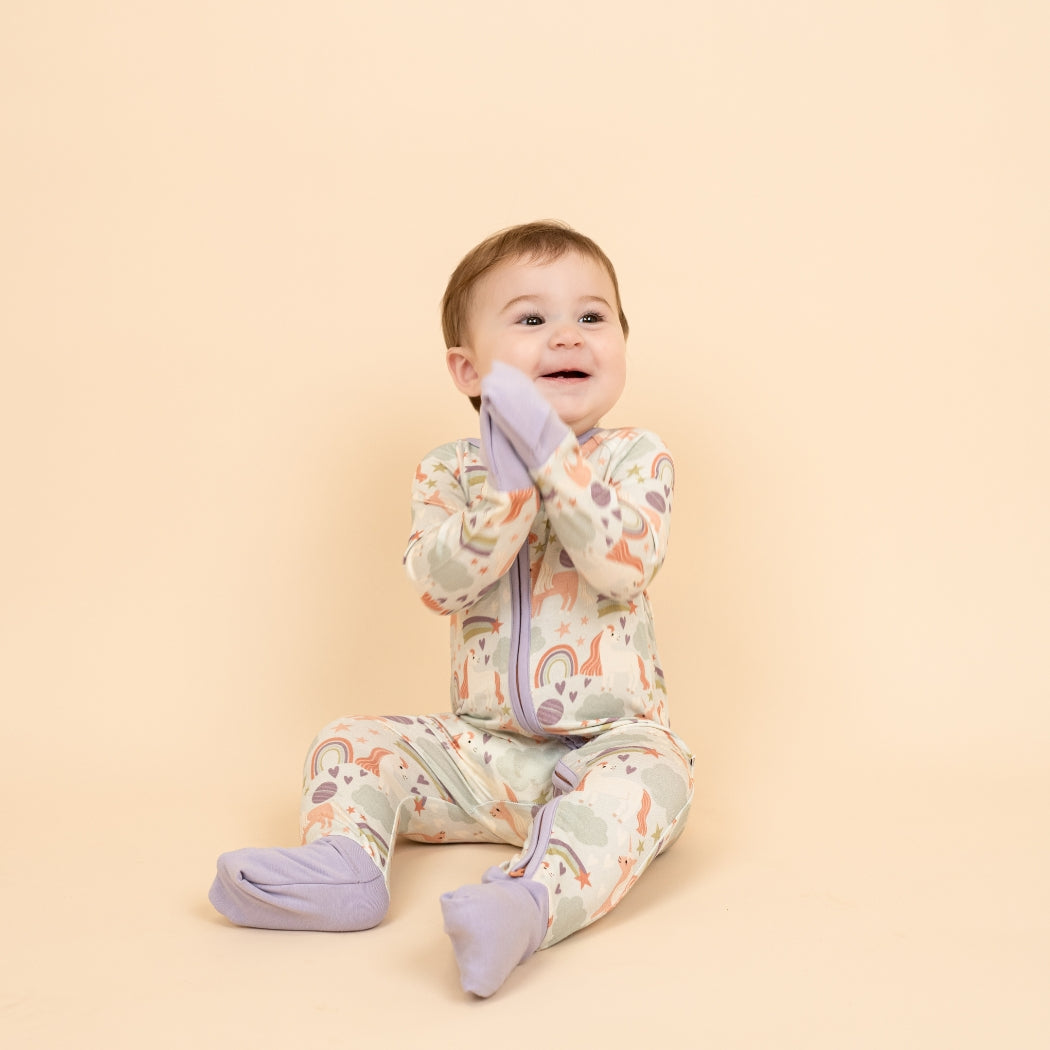 Excited little girl in our Magical Unicorn bamboo convertible pajama clapping hands together. Her feet and hands have the foldable covers on them from the pajama. The print includes unicorns, rainbows, clouds, planets, stars, and hearts. The colors are purple, pink, and blu, with hints of green.