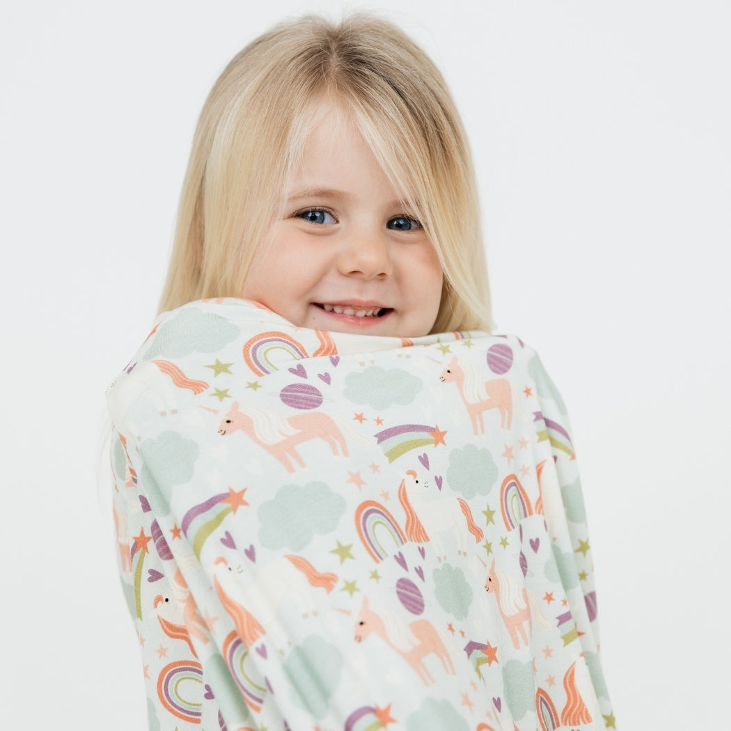 Little girl wearing her Magical Unicorn pajama set, wrapped up in our Magical Unicorn blanket. The print includes unicorns, rainbows, clouds, planets, stars, and hearts. The colors are purple, pink, and blu, with hints of green.