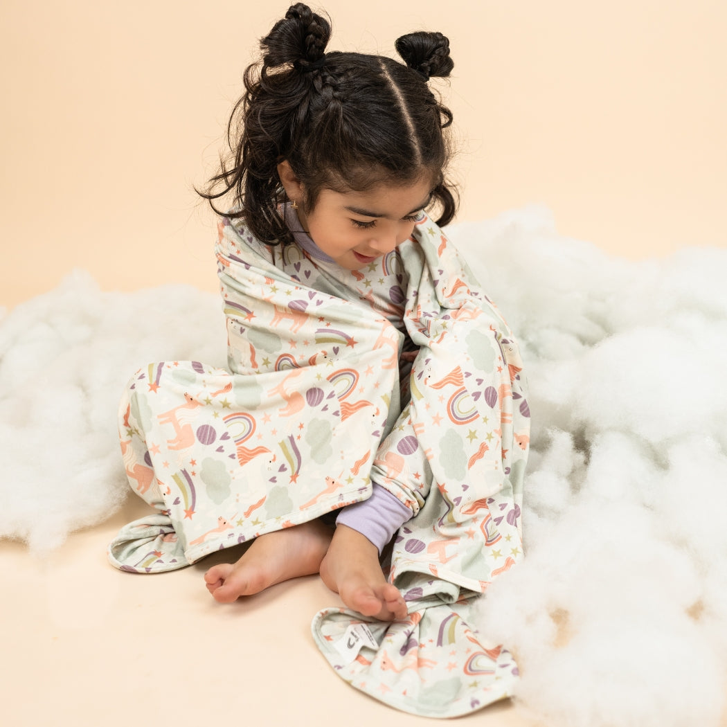 Little girl atop fake clouds wearing her Magical Unicorn pajama set, wrapped up in our Magical Unicorn blanket. The print includes unicorns, rainbows, clouds, planets, stars, and hearts. The colors are purple, pink, and blu, with hints of green.