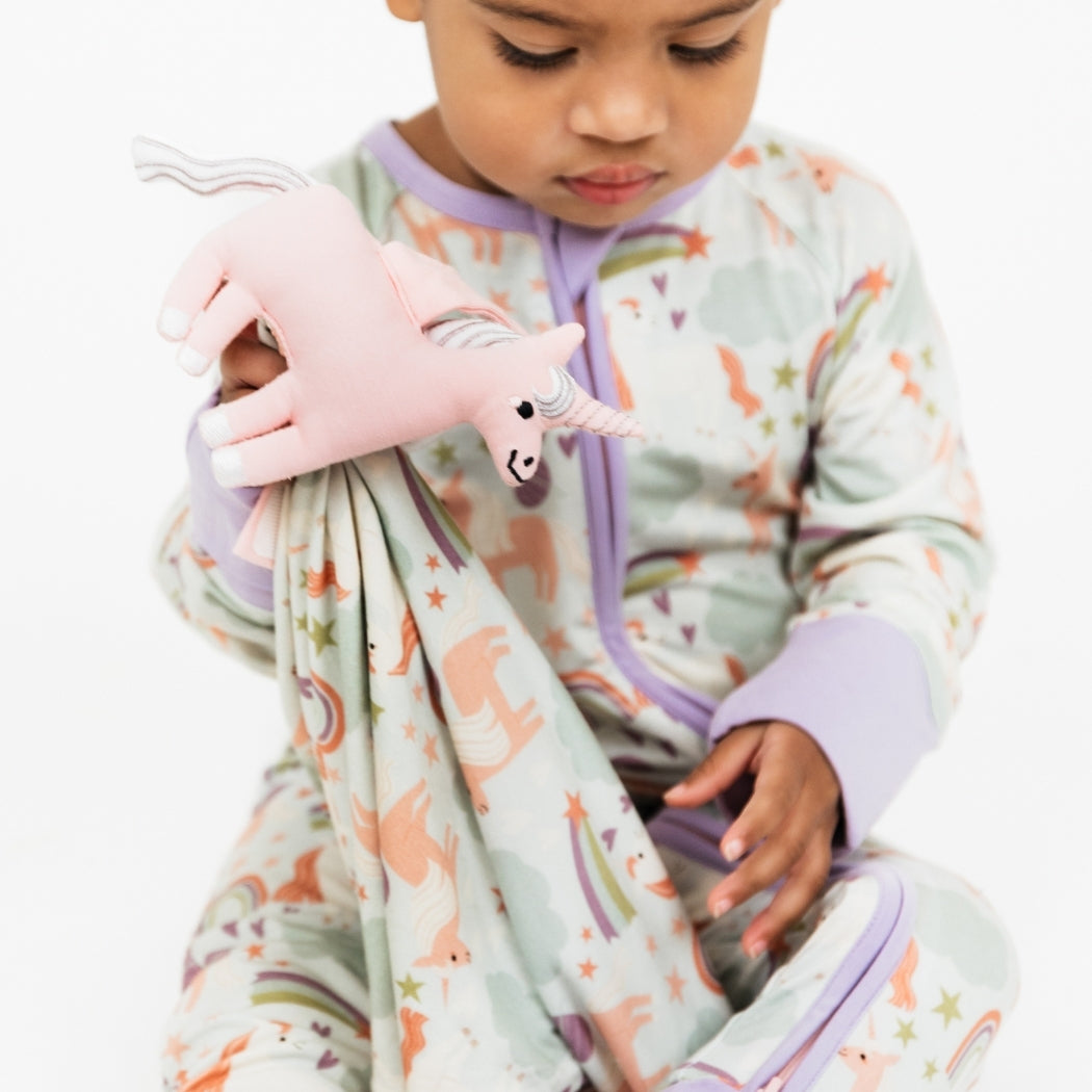 Little girl wearing our Magical Unicorn convertible holding the Magical Unicorn Lovey. The print includes unicorns, rainbows, clouds, planets, stars, and hearts. The colors are purple, pink, and blu, with hints of green.