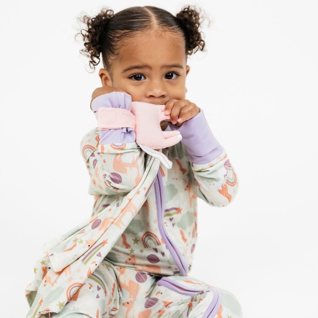 Little girl wearing our Magical Unicorn convertible holding the Magical Unicorn Lovey. The print includes unicorns, rainbows, clouds, planets, stars, and hearts. The colors are purple, pink, and blu, with hints of green.