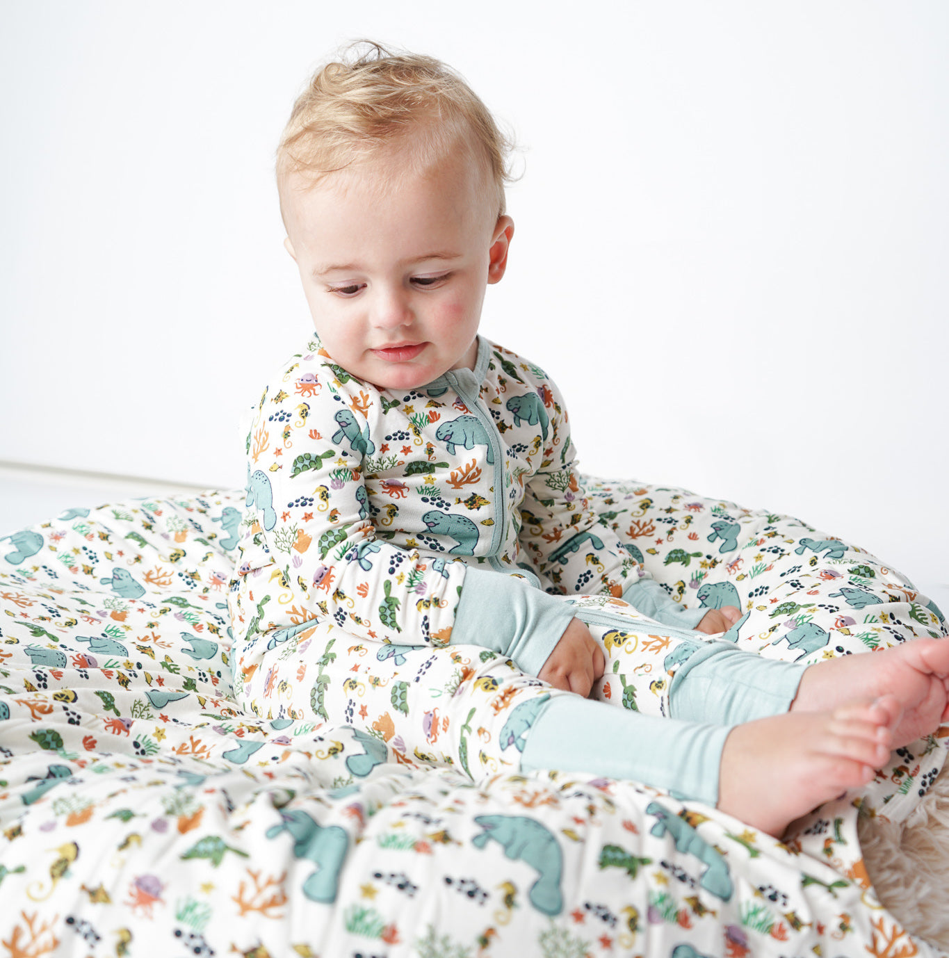 toddler sit on the "manatee" bamboo blanket, wearing the "manatee" convertible. the print "manatee" unites the florida sea life in a majestic print. there is a variation of florida manatees, yellow and pink sea horses, yellow and pink starfish, bubbles, turtles, and coral patterned across a white background.