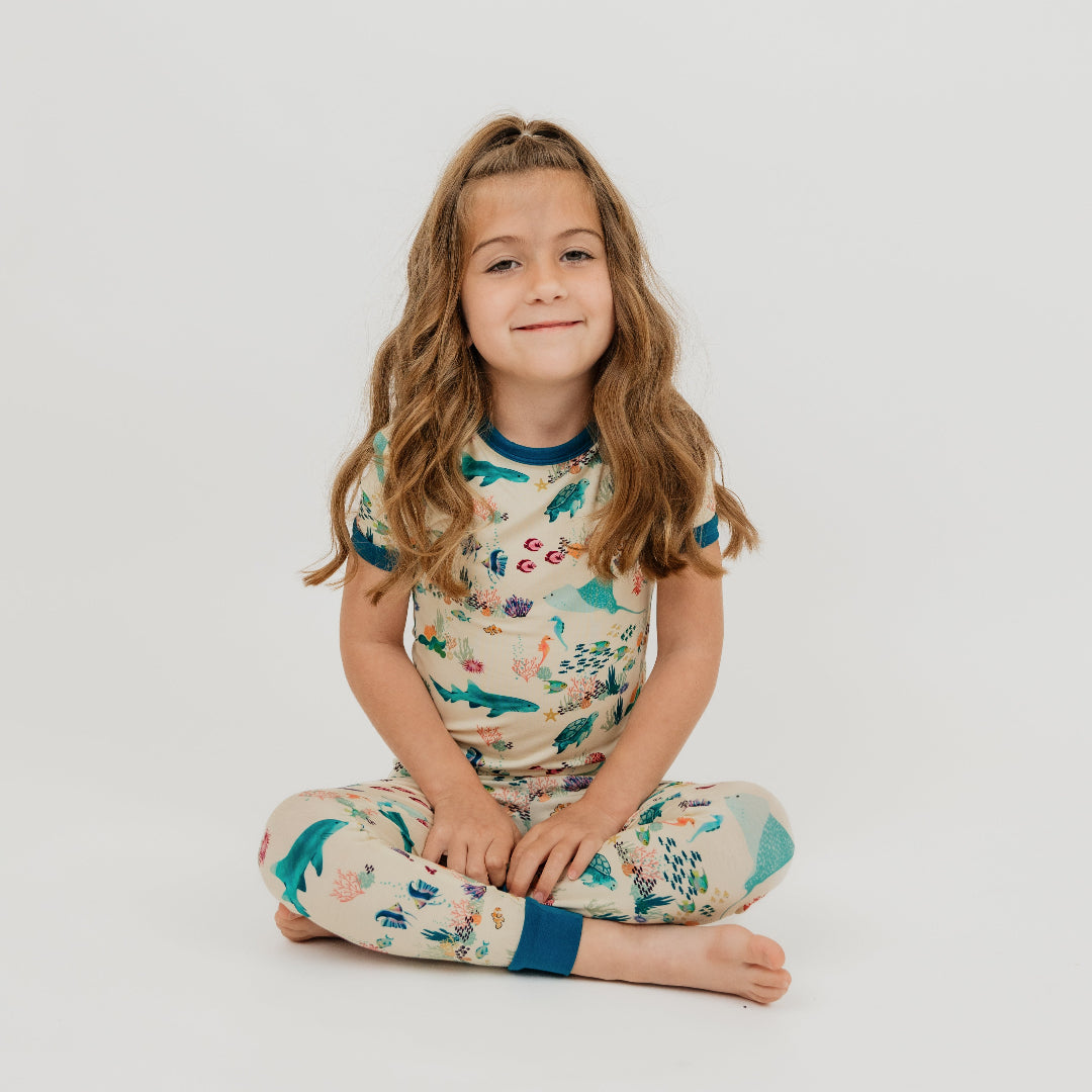 Toddler girl sitting down in a white background wearing our Emerson and Friends Coral Reef Bamboo Kids Pajama Set featuring a soft, breathable short sleeve top and matching pants with an ocean-inspired print of sharks, sea turtles, and coral reefs—perfect for cozy nights and sweet dreams.