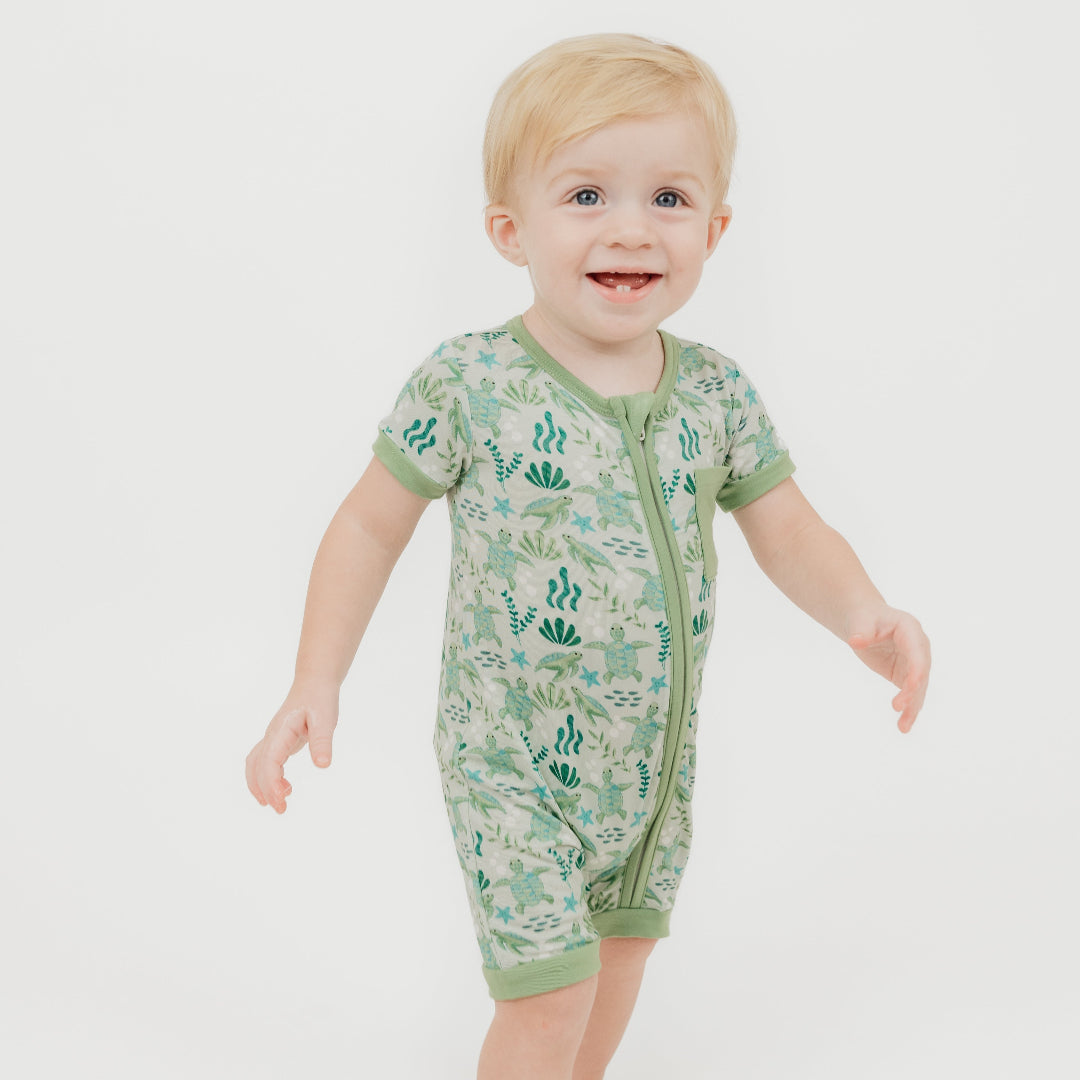 A toddler enjoying our Save the Sea Turtle shortie romper, made from breathable, eco-friendly cotton. This soft and breathable short-sleeve romper features a charming ocean-inspired turtle print, a front zipper for easy changes, and a cozy pocket detail.