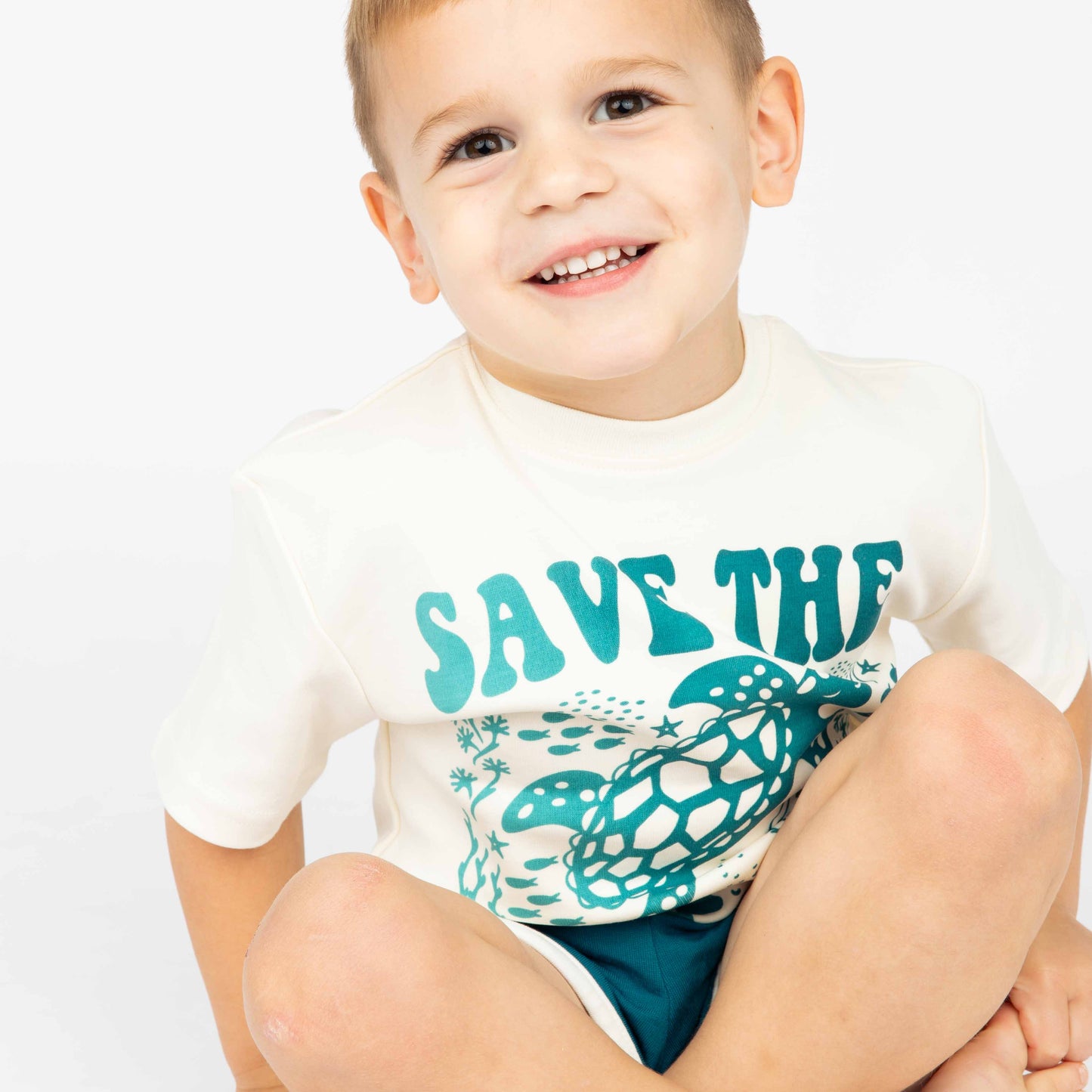 A boy smiling wearing our Save the Sea Turtle Tee while sitting down in a white background.