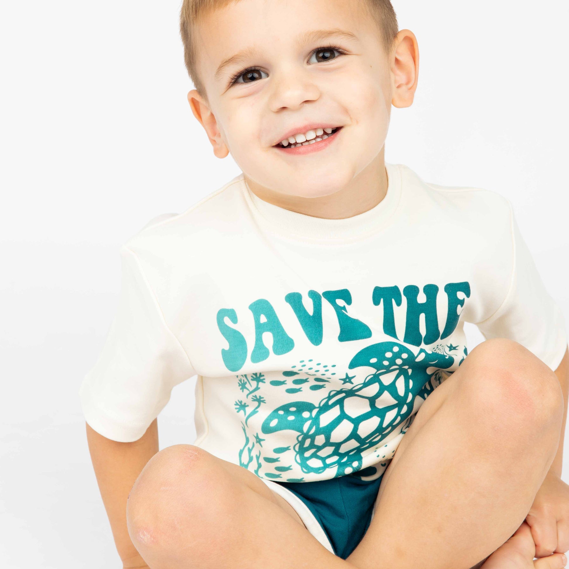 A boy smiling wearing our Save the Sea Turtle Tee while sitting down in a white background.