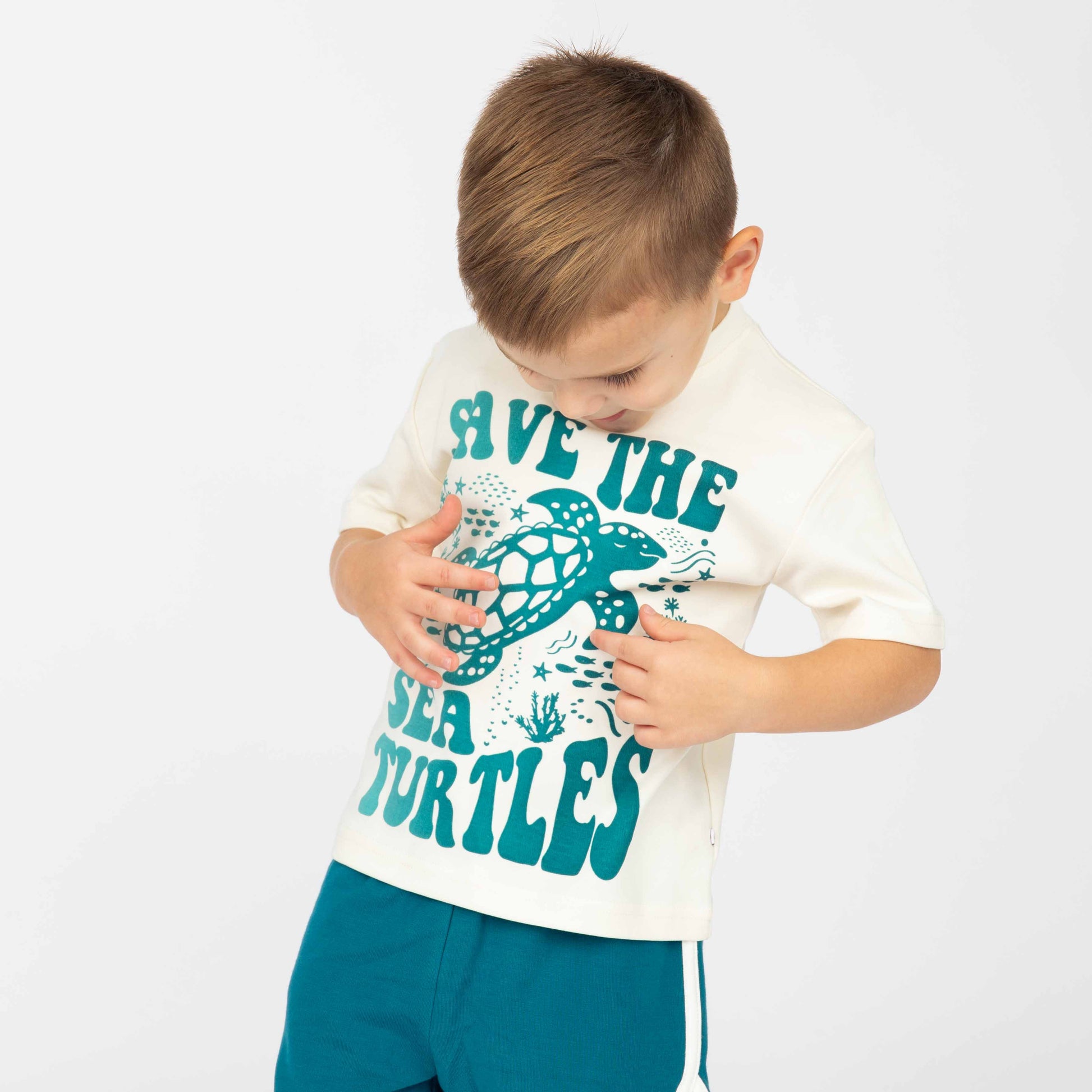 The boy is pointing at the Save the Sea Turtle T-shirt showing us the turtle. Modeling in a white background.