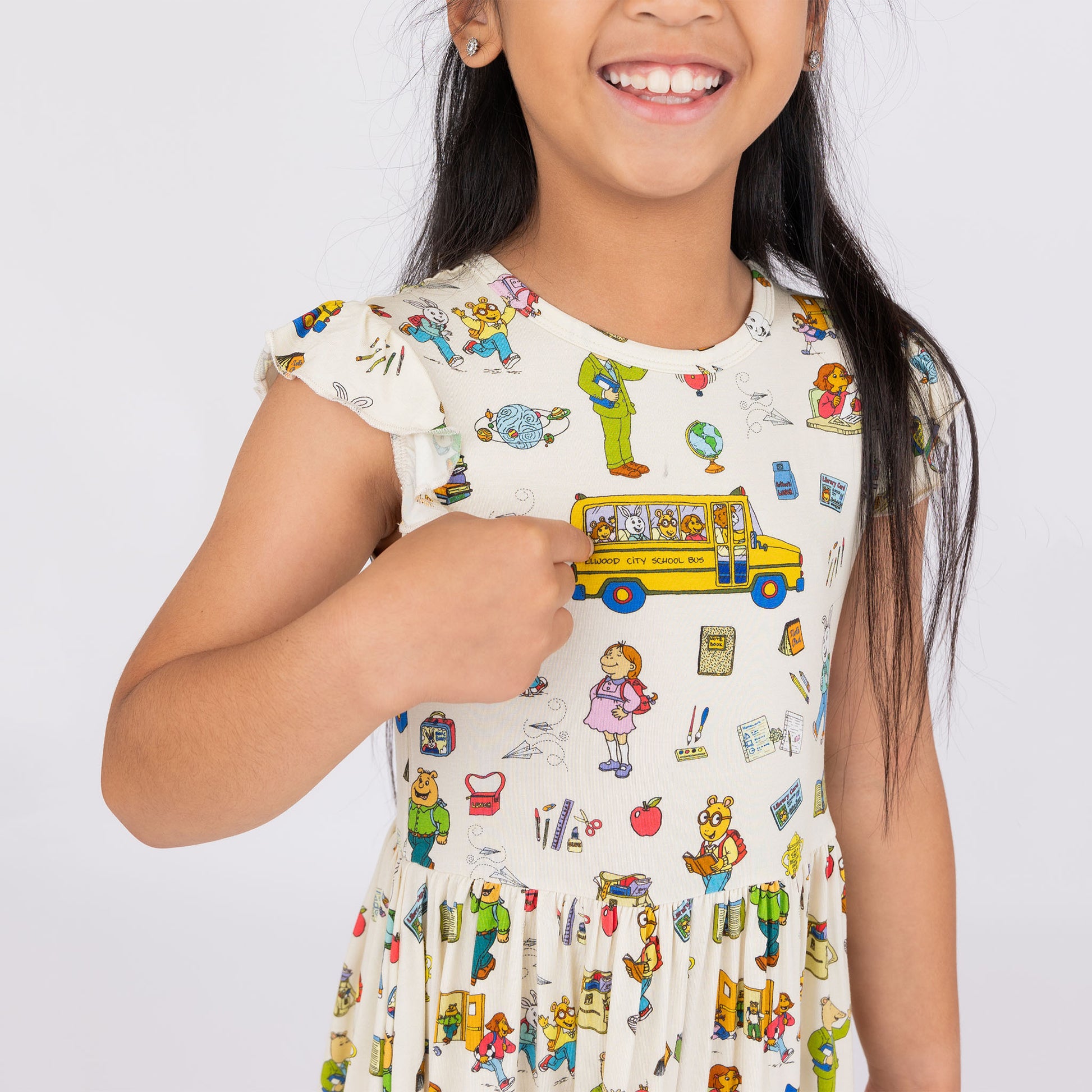 Girl pointing at the School Bus in our twirl bamboo dress featuring playful illustrations of Arthur™ PBS Character, books, and school bus, a beige background.