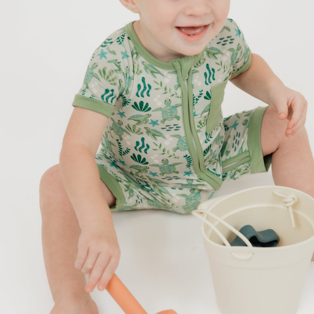 A toddler sitting down enjoying our Save the Sea Turtle shortie romper, made from breathable, eco-friendly cotton. This soft and breathable short-sleeve romper features a charming ocean-inspired turtle print, a front zipper for easy changes, and a cozy pocket detail.