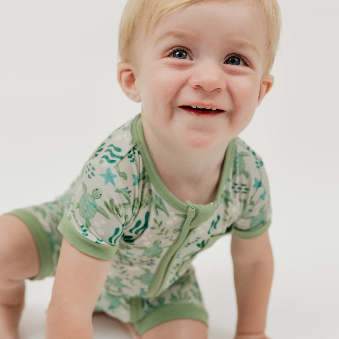 A smiling toddler enjoying our Save the Sea Turtle shortie romper, made from breathable, eco-friendly cotton. This soft and breathable short-sleeve romper features a charming ocean-inspired turtle print, a front zipper for easy changes, and a cozy pocket detail.