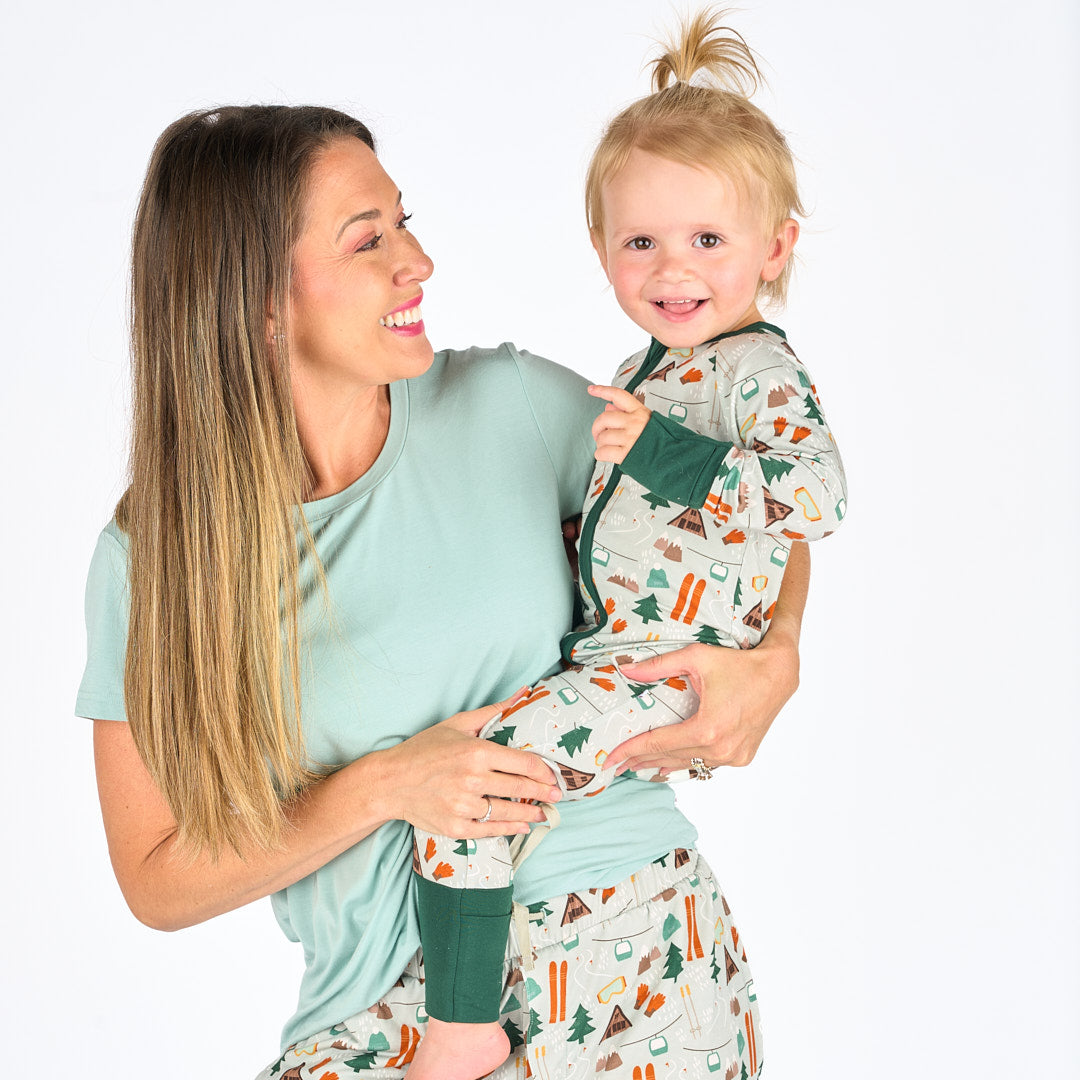 Mother and daughter modeling in white background wearing bamboo pajama ski day featuring trees, cabins, and other ski activities.