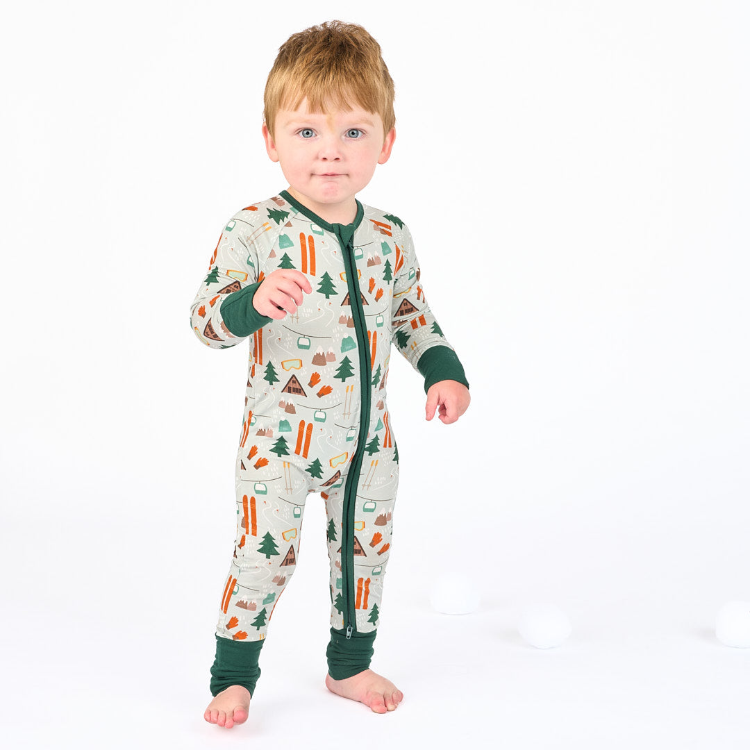 toddler modeling in white background wearing bamboo pajama ski day featuring trees, cabins, and other ski activities.