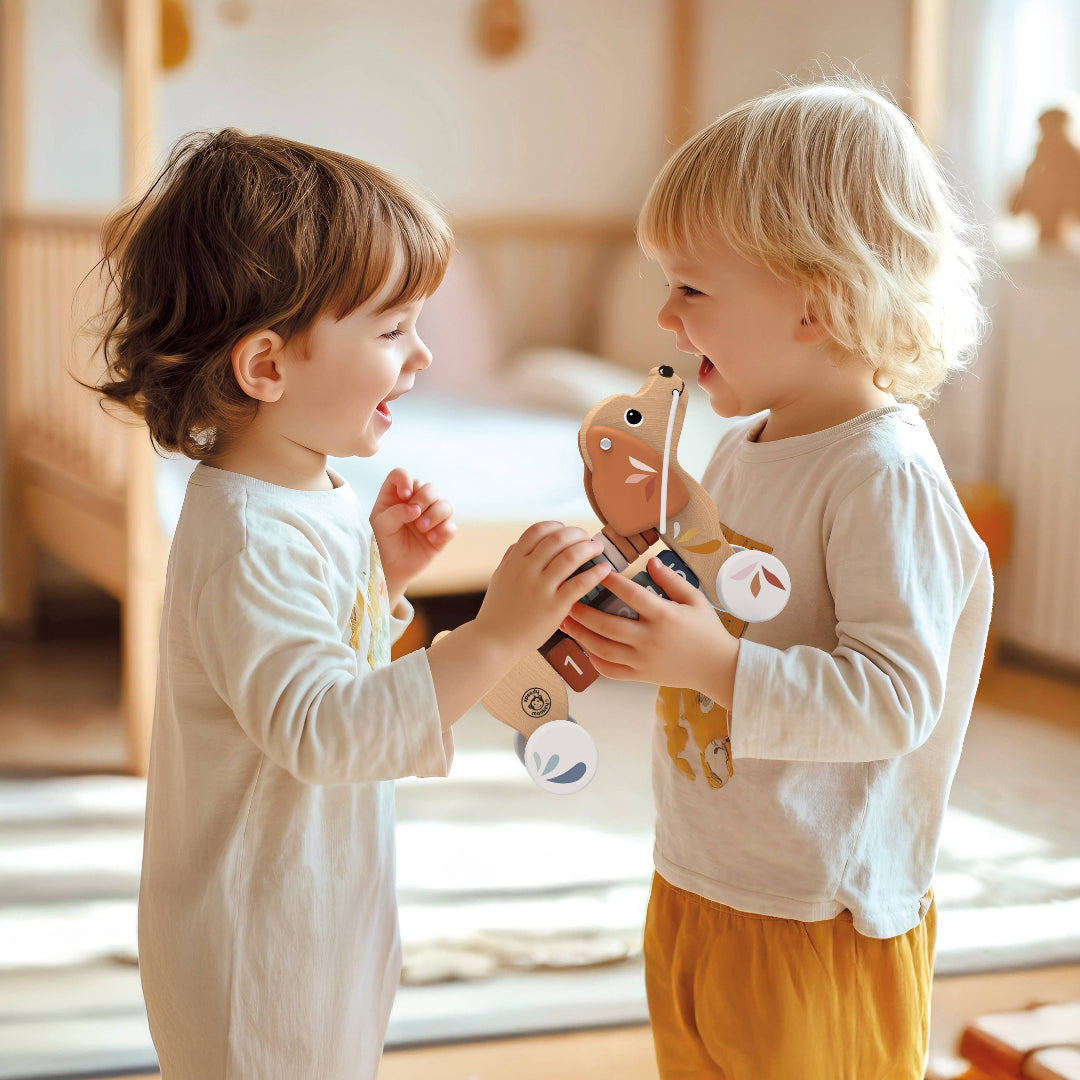 2 toddlers playing with a wooden pull along dog toy in a bedroom with a bed in the background