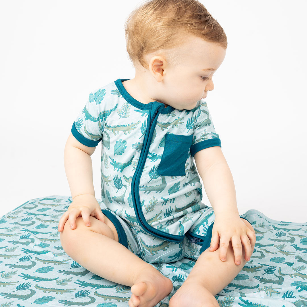 little toddler boy sitting on the "later gator" blanket. he is wearing the "later gator" shortie. the "later gator" print has a mix of light and dark green alligators, leaves, and white dots scattered on a teal colored background.