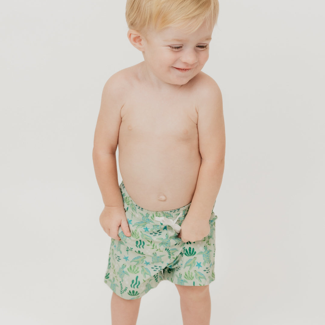 Smiling boy wearing our Save the Sea Turtle Swim trunks. The print is featuring a sea-green turtle and ocean-inspired print. Made from quick-dry, lightweight fabric with an adjustable drawstring waist for a comfy fit. Perfect for beach days and poolside fun!