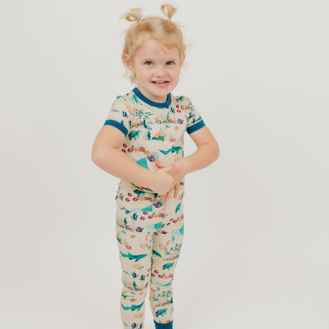 Toddler girl wearing Emerson and Friends Coral Reef Bamboo Kids Pajama Set featuring a soft, breathable short sleeve top and matching pants with an ocean-inspired print of sharks, sea turtles, and coral reefs—perfect for cozy nights and sweet dreams.