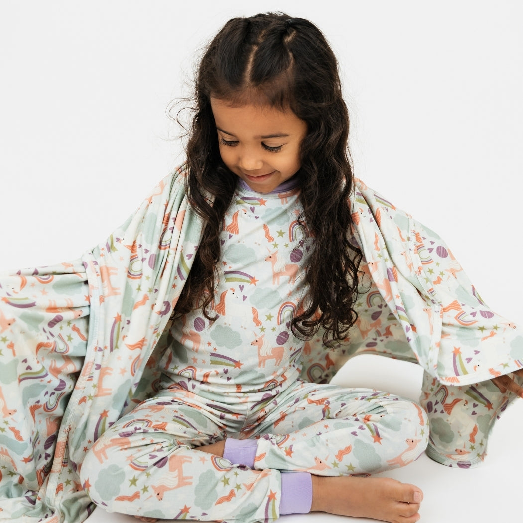 Little girl wearing her Magical Unicorn pajama set, wrapped up in our Magical Unicorn blanket. The print includes unicorns, rainbows, clouds, planets, stars, and hearts. The colors are purple, pink, and blu, with hints of green.