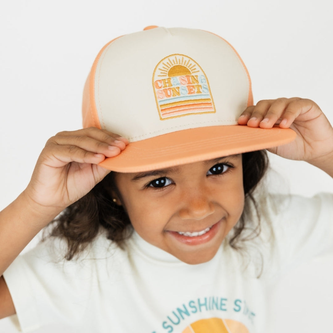 Little girl wearing our Chasing Sunsets Snapback hat holding onto the rim and smiling up at the camera.