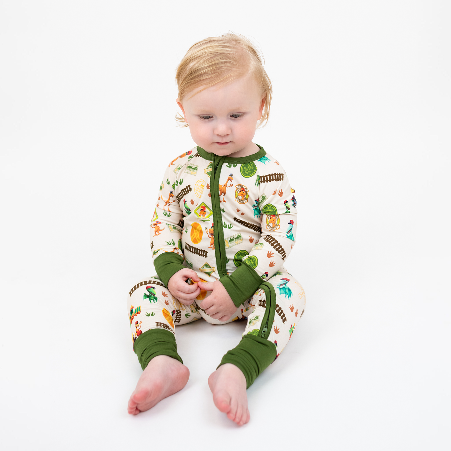 Toddler sitting down with our Emerson and Friends All Aboard the Dino Train bamboo convertible baby pajamas with green trim, double zipper, and colorful orange, blue, and green dinosaurs on train tracks with "Dino Train" print.