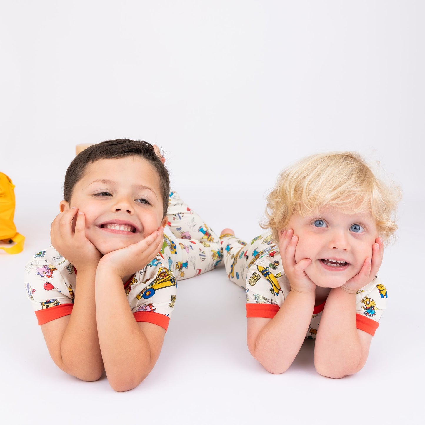 Siblings wearing the short Sleeve Kids Pajama Pants Set from buttery-soft bamboo, featuring red trims and playful illustrations of Arthur™ PBS Character, books, and school bus, a beige background.