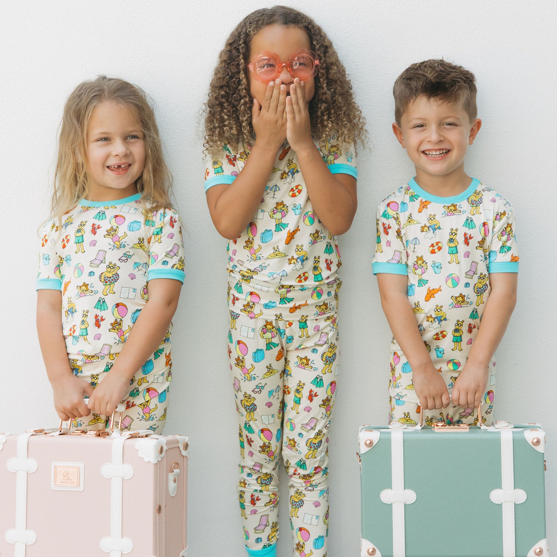 Siblings wearing our Arthur's Family Vacation Bamboo Short Sleeve Kids Pajama Pants Set featuring vibrant light blue trim and a playful print of Arthur™ in swimwear surrounded by fun beach props — perfect for family getaways.