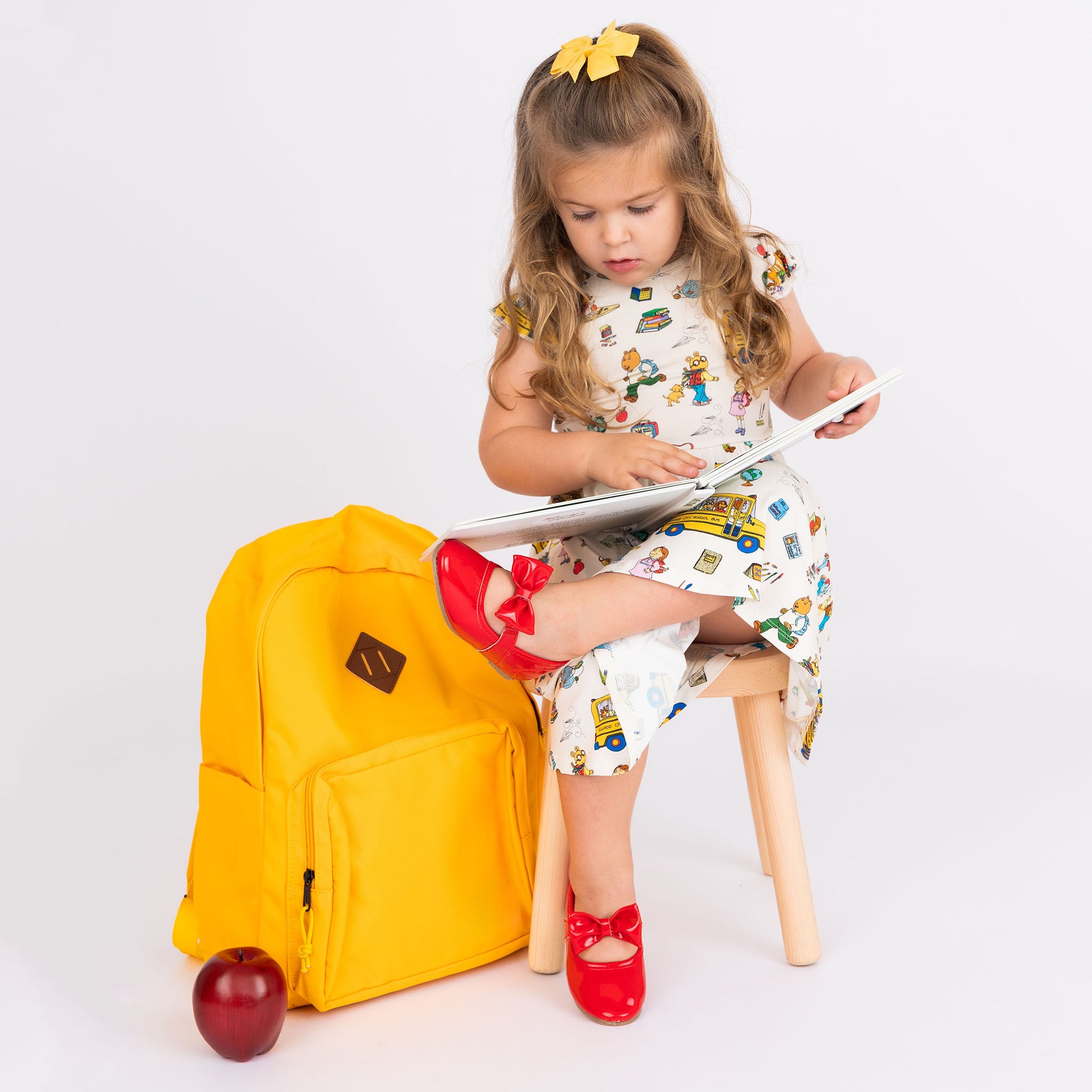 Girl sitting in a wooden char reading and wearing our twirl bamboo dress featuring playful illustrations of Arthur™ PBS Character, books, and school bus, a beige background.