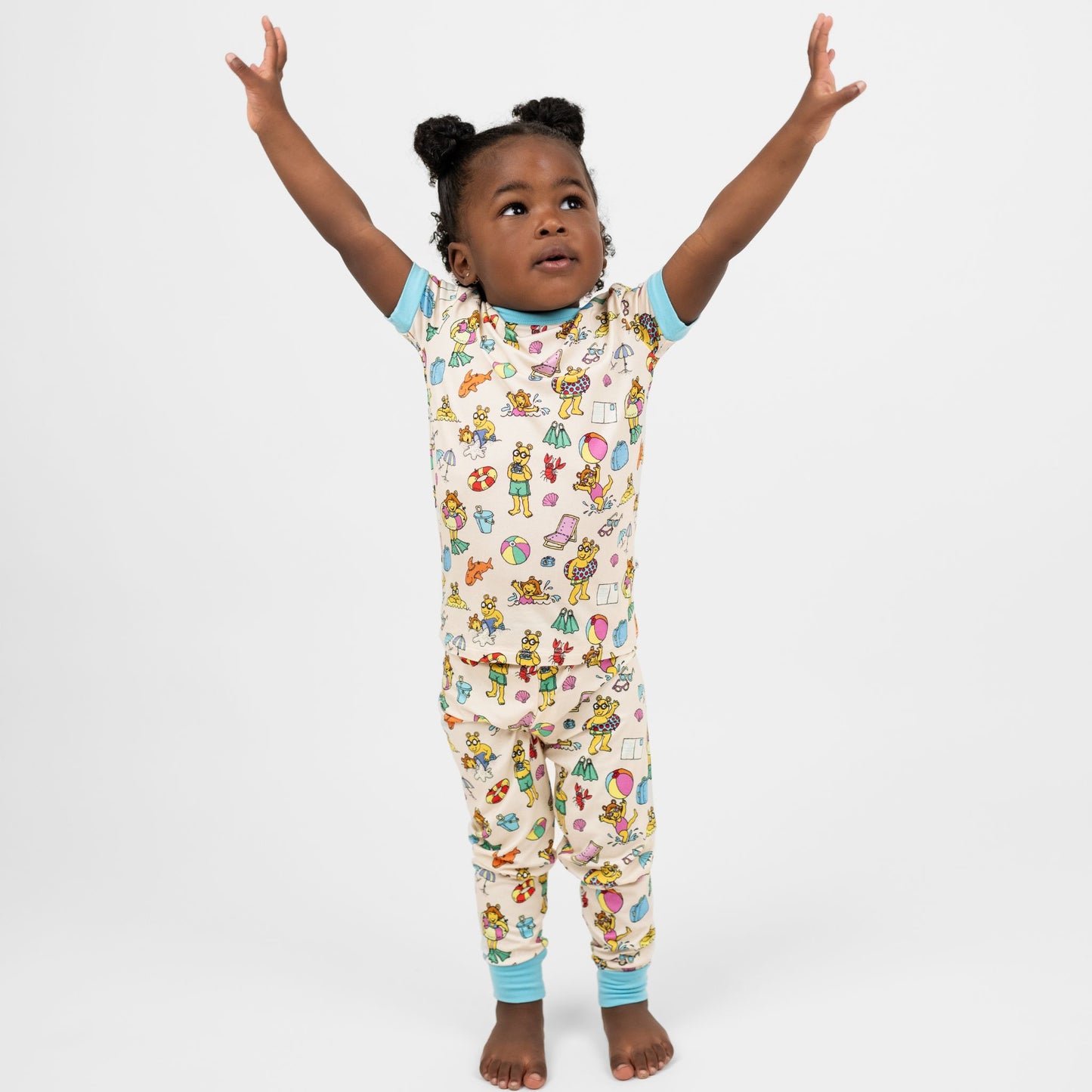 Toddler wearing our Arthur's Family Vacation Bamboo Short Sleeve Kids Pajama Pants Set featuring vibrant light blue trim and a playful print of Arthur™ in swimwear surrounded by fun beach props — perfect for family getaways.