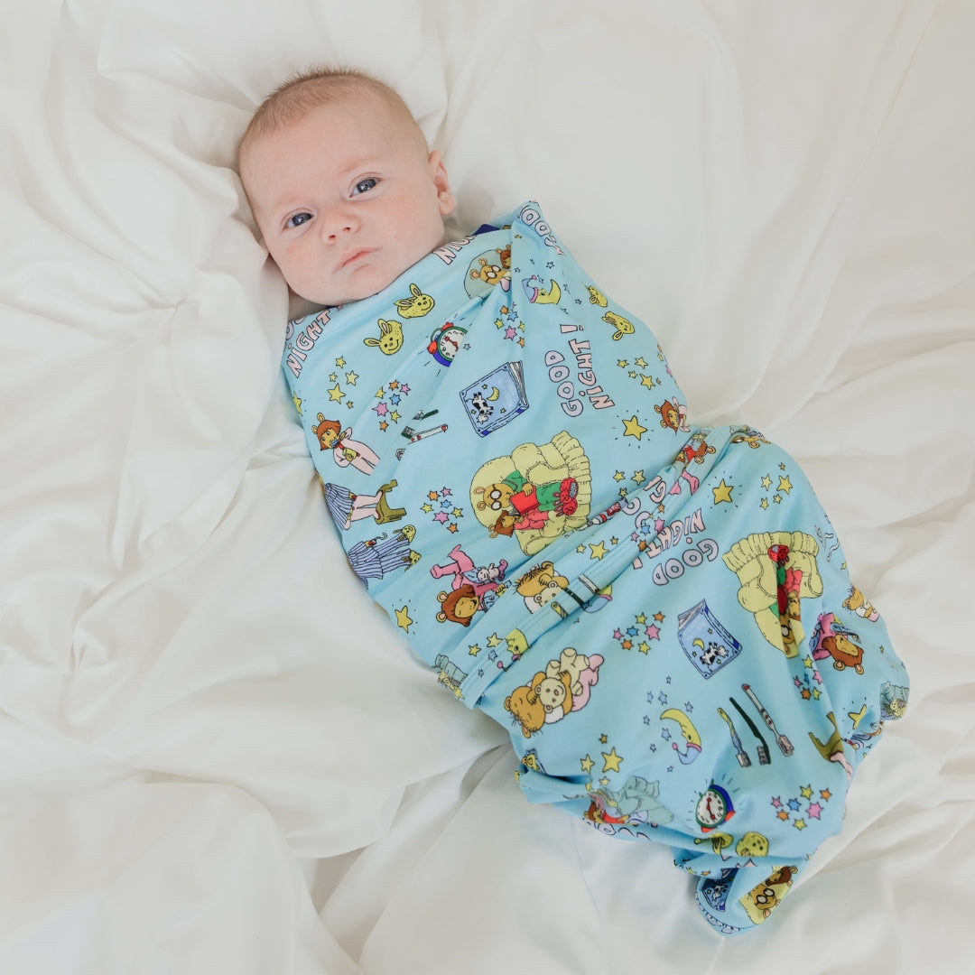 Baby swaddled in our PBS Good Night Arthur™ collaboration blanket with Arthur and friends in a cozy bedtime scene complete with storybooks, toothbrushes, bunny slippers, a sleeping puppy, crescent moons with sleep caps, twinkling stars, and the words "GOOD NIGHT!" on a soft blue background.