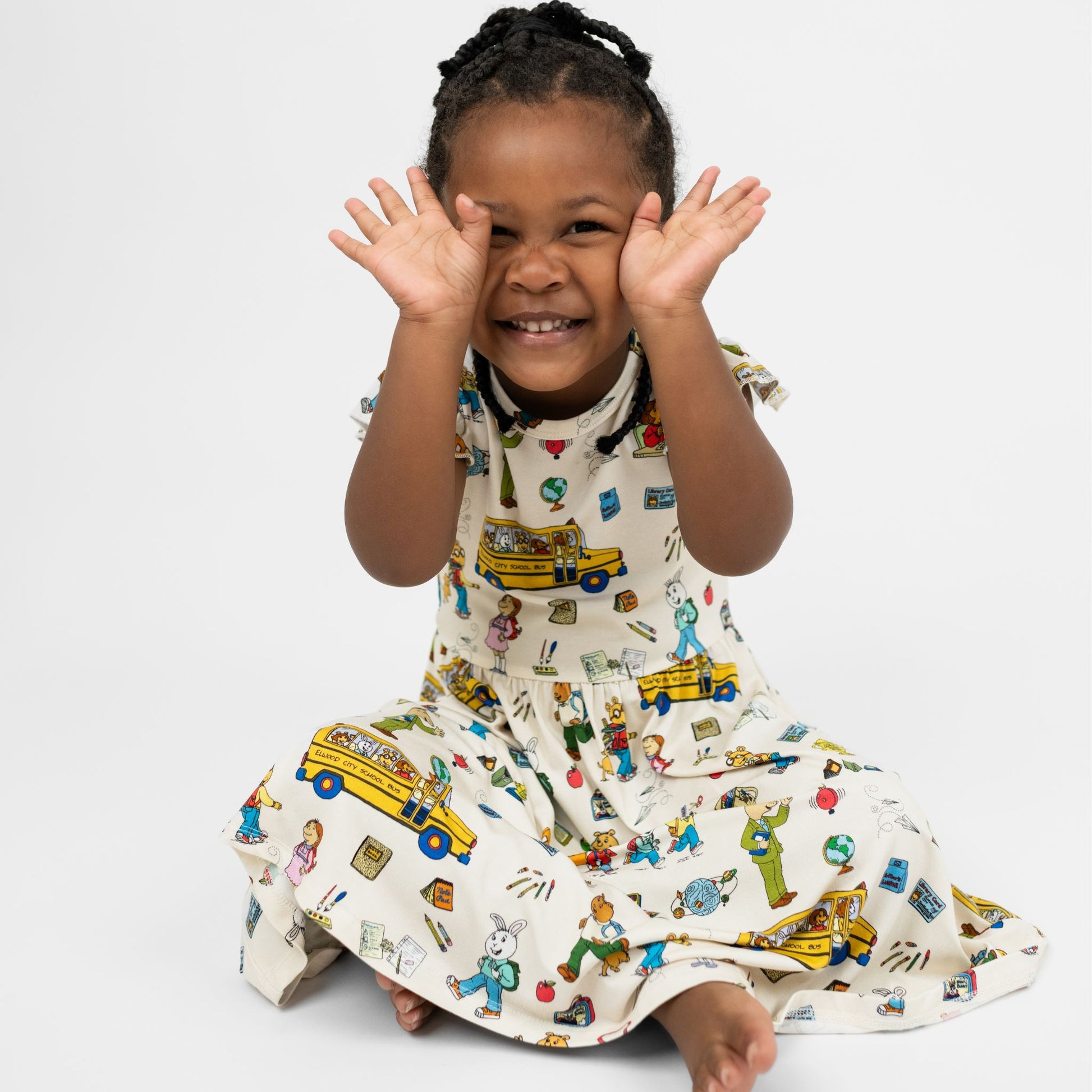 Toddler girl sitting on the floor wearing our twirl bamboo dress featuring playful illustrations of Arthur™ PBS Character, books, and school bus, a beige background.