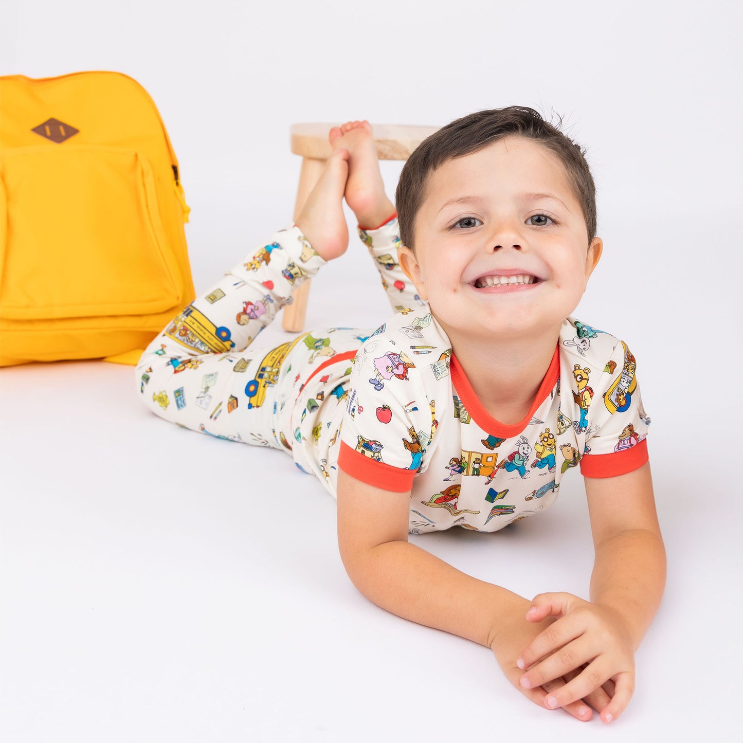 Toddler boy laying down in the floor smiling wearing our Short Sleeve Kids Pajama Pants Set from buttery-soft bamboo, featuring red trims and playful illustrations of Arthur™ PBS Character, books, and school bus, a beige background.