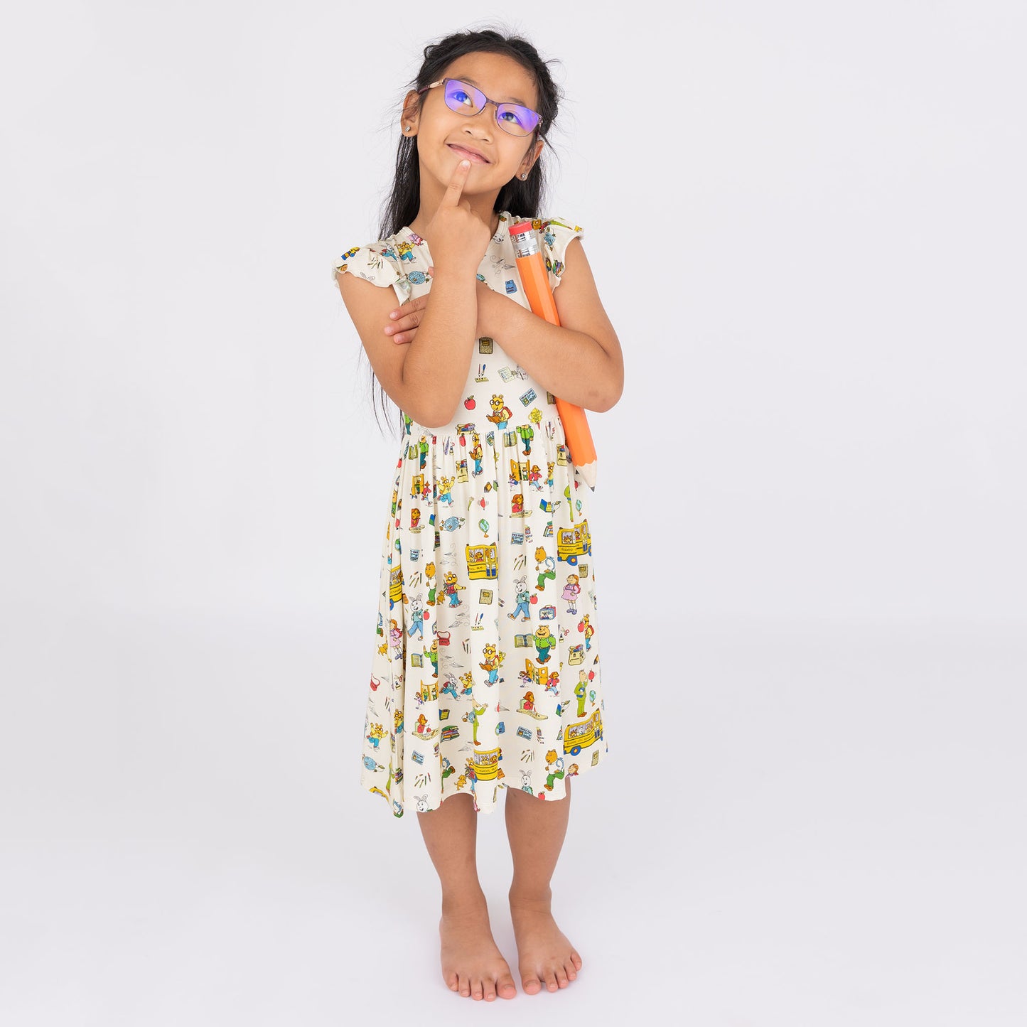 Girl wearing our twirl bamboo dress featuring playful illustrations of Arthur™ PBS Character, books, and school bus, a beige background.