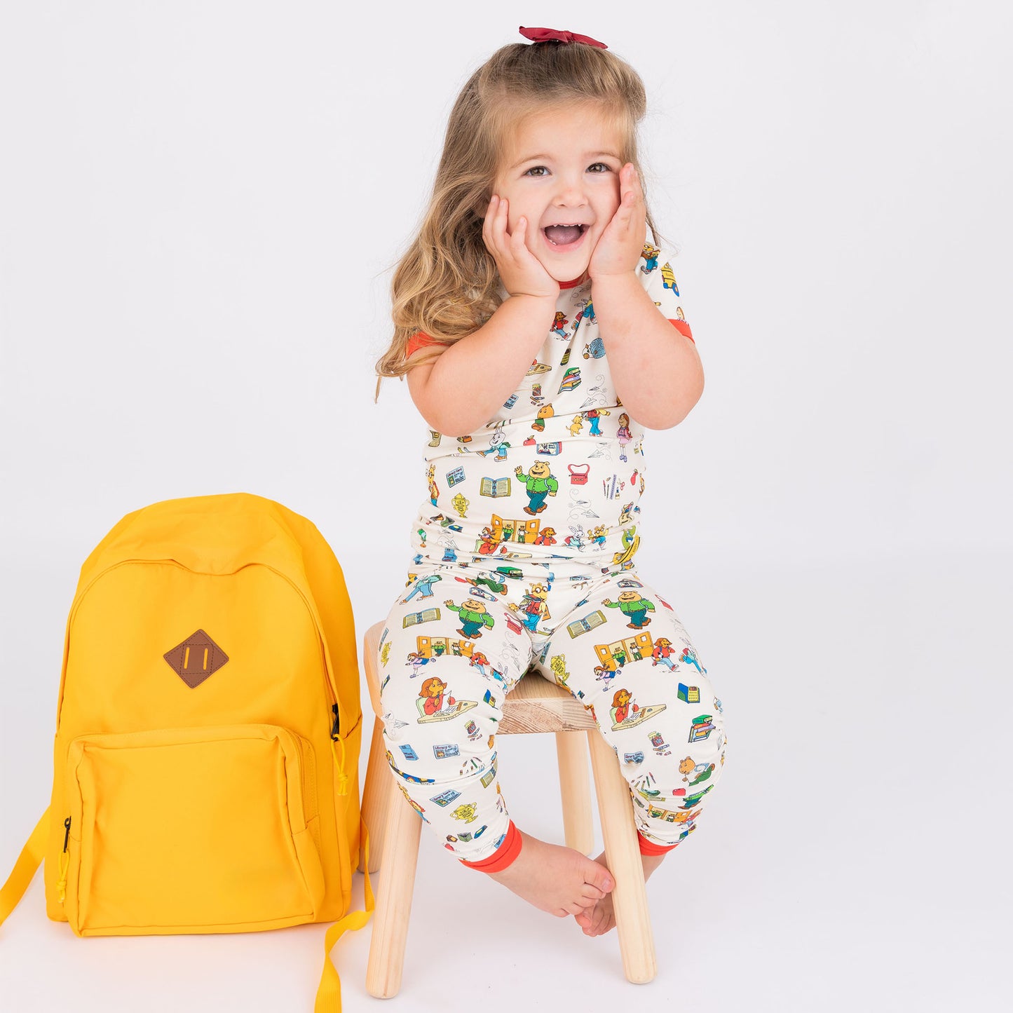Girl smiling at the camera with a yellow backpack on the side with out short Sleeve Kids Pajama Pants Set from buttery-soft bamboo, featuring red trims and playful illustrations of Arthur™ PBS Character, books, and school bus, a beige background.