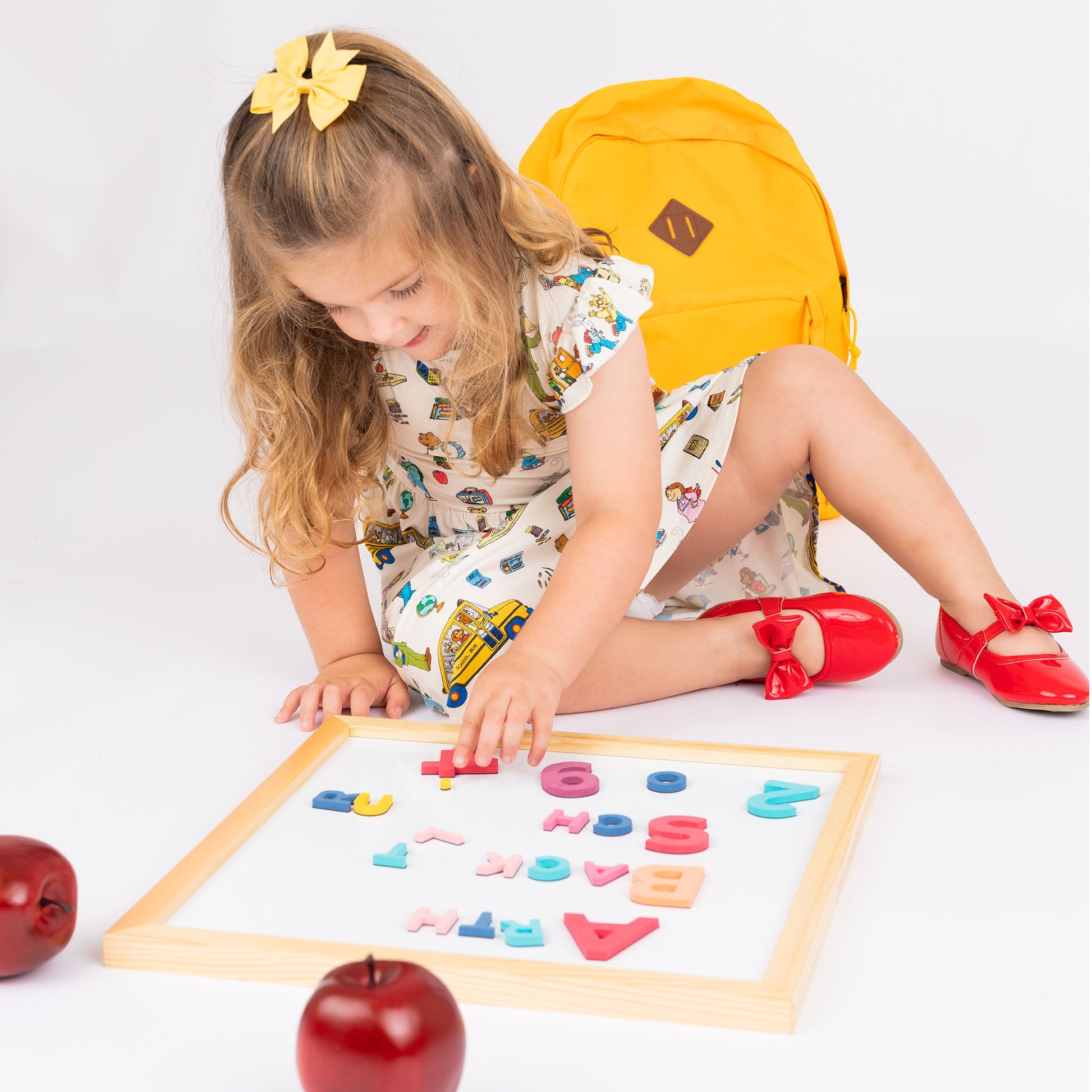Girl sitting down playing with letter and magnetic white board wearing our twirl bamboo dress featuring playful illustrations of Arthur™ PBS Character, books, and school bus, a beige background.