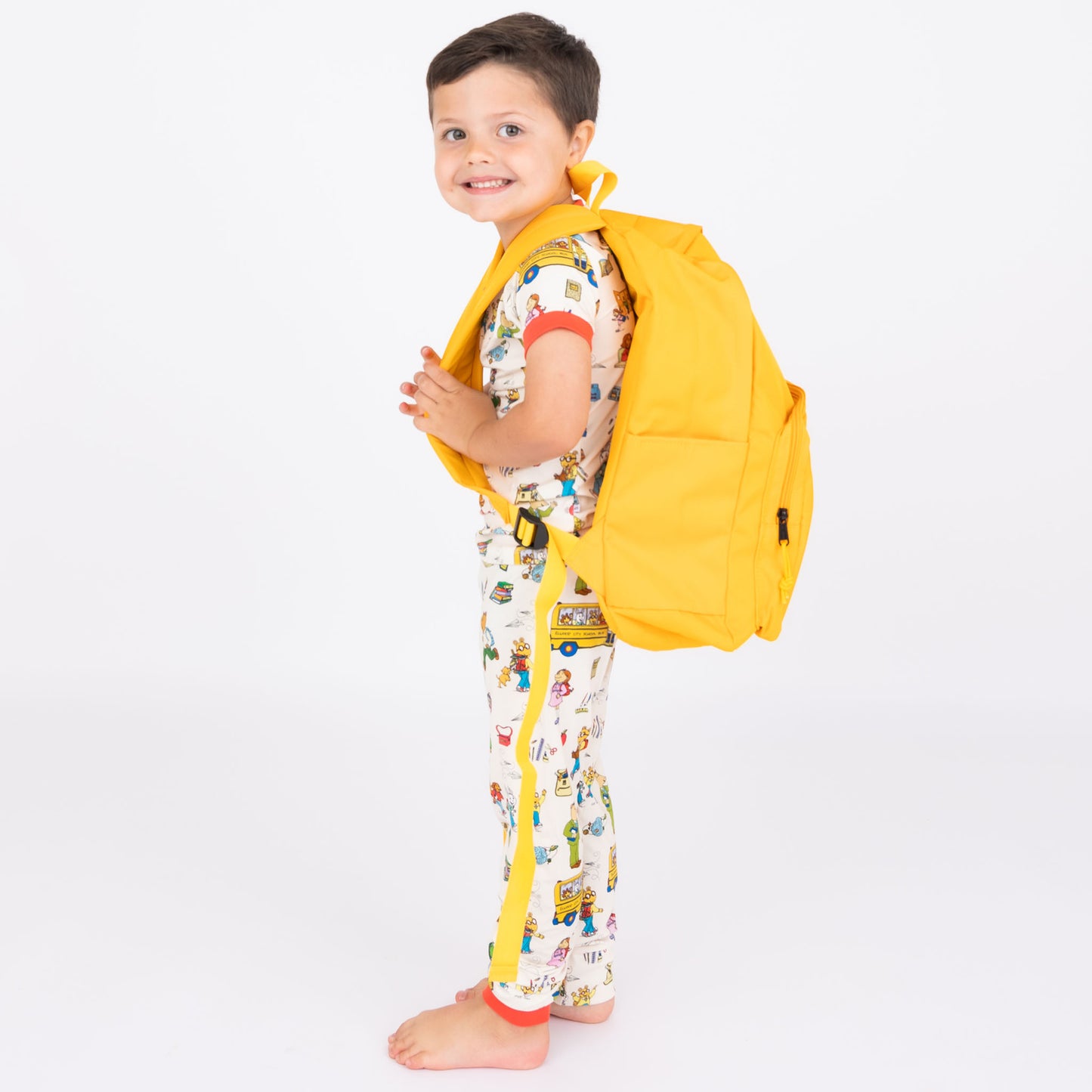 Boy wearing our short Sleeve Kids Pajama Pants Set from buttery-soft bamboo, featuring red trims and playful illustrations of Arthur™ PBS Character, books, and school bus, a beige background.