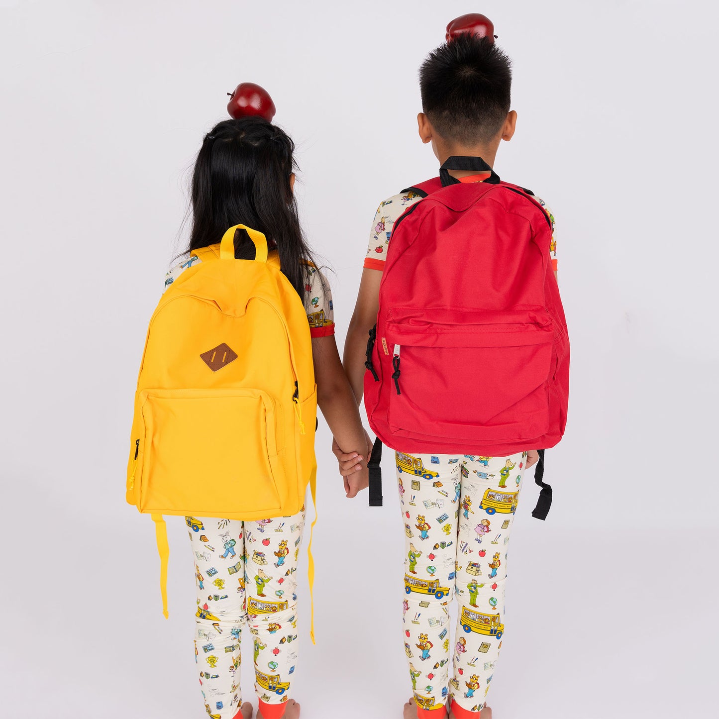 Siblings wearing the short Sleeve Kids Pajama Pants Set from buttery-soft bamboo, featuring red trims and playful illustrations of Arthur™ PBS Character, books, and school bus, a beige background.