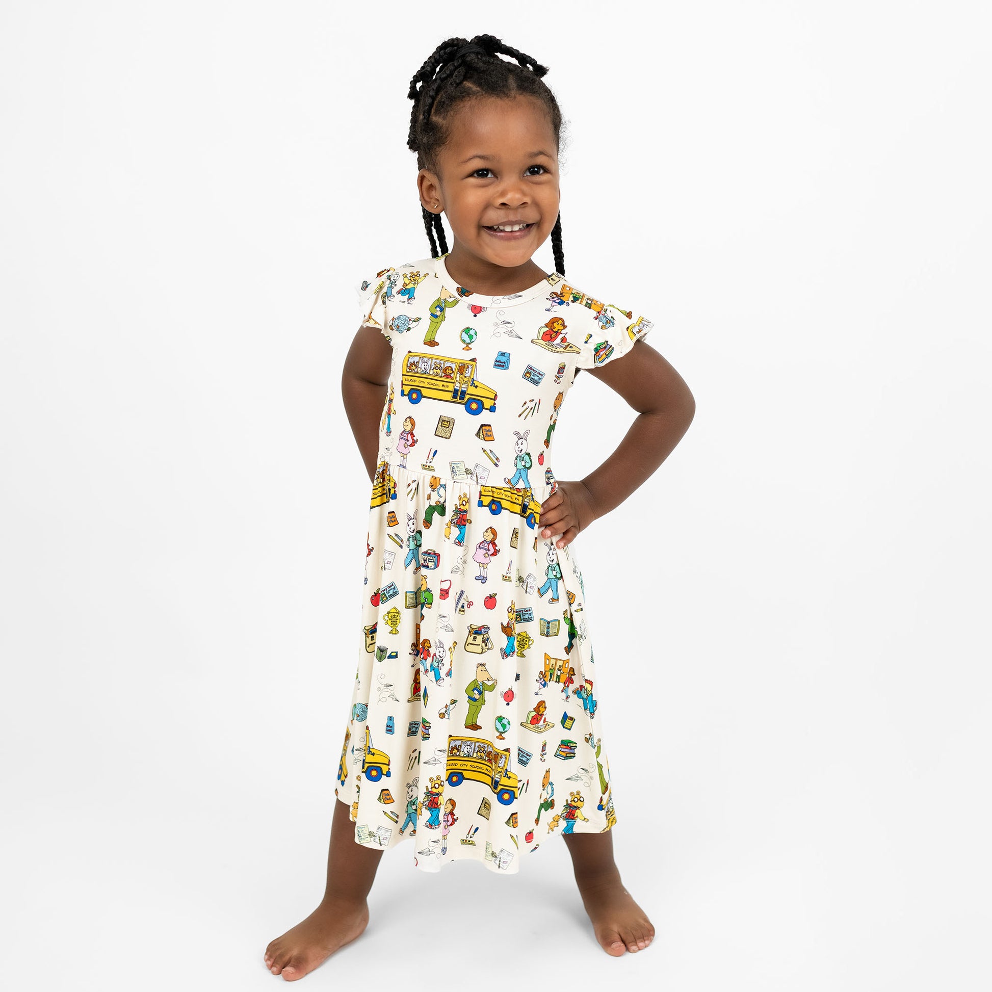 Toddler girl happy wearing our twirl bamboo dress featuring playful illustrations of Arthur™ PBS Character, books, and school bus, a beige background.