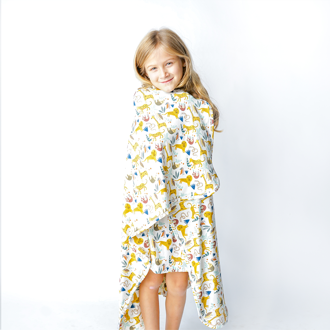 a girl wraps herself in the "jungle friends" bamboo blanket. the "jungle friends" print is a mix of jungle animals that include giraffes, lions, cheetahs, sloths, snakes, and forest plants.