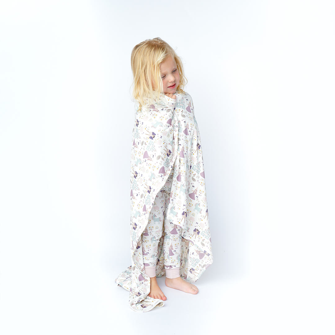 a young girl wraps herself in the "once upon a time" bamboo blanket. the "once upon a time" print is a mix of pinks, purples, whites, blues, and yellows. you can see fairy princess and regular princess, unicorns, swans, and castles. theres also hearts, stars and sparkles scattered around the print.