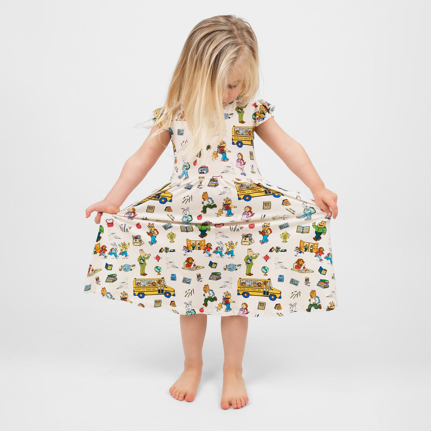 Toddler girl sitting in a chair wearing our twirl bamboo dress featuring playful illustrations of Arthur™ PBS Character, books, and school bus, a beige background.