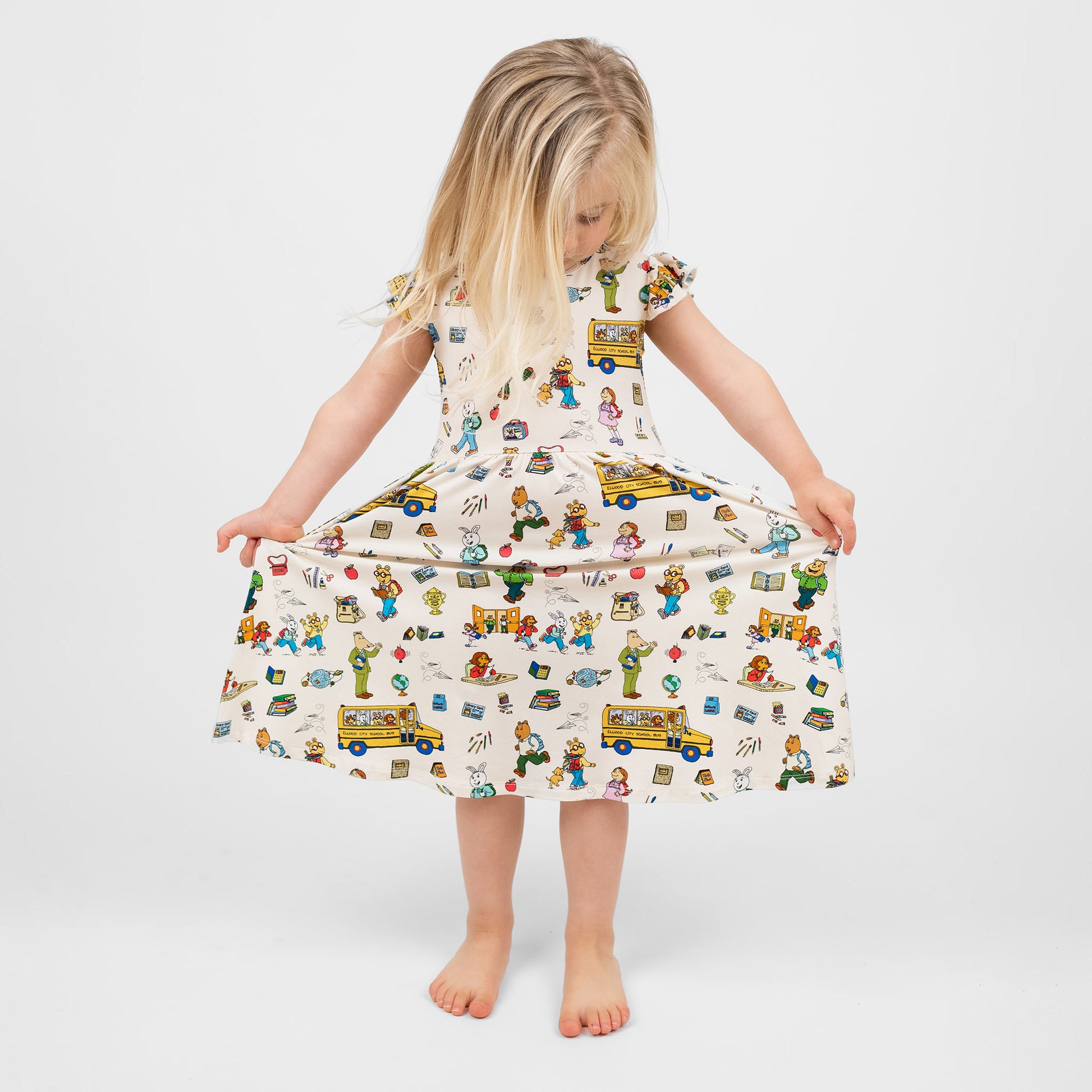Toddler girl sitting in a chair wearing our twirl bamboo dress featuring playful illustrations of Arthur™ PBS Character, books, and school bus, a beige background.