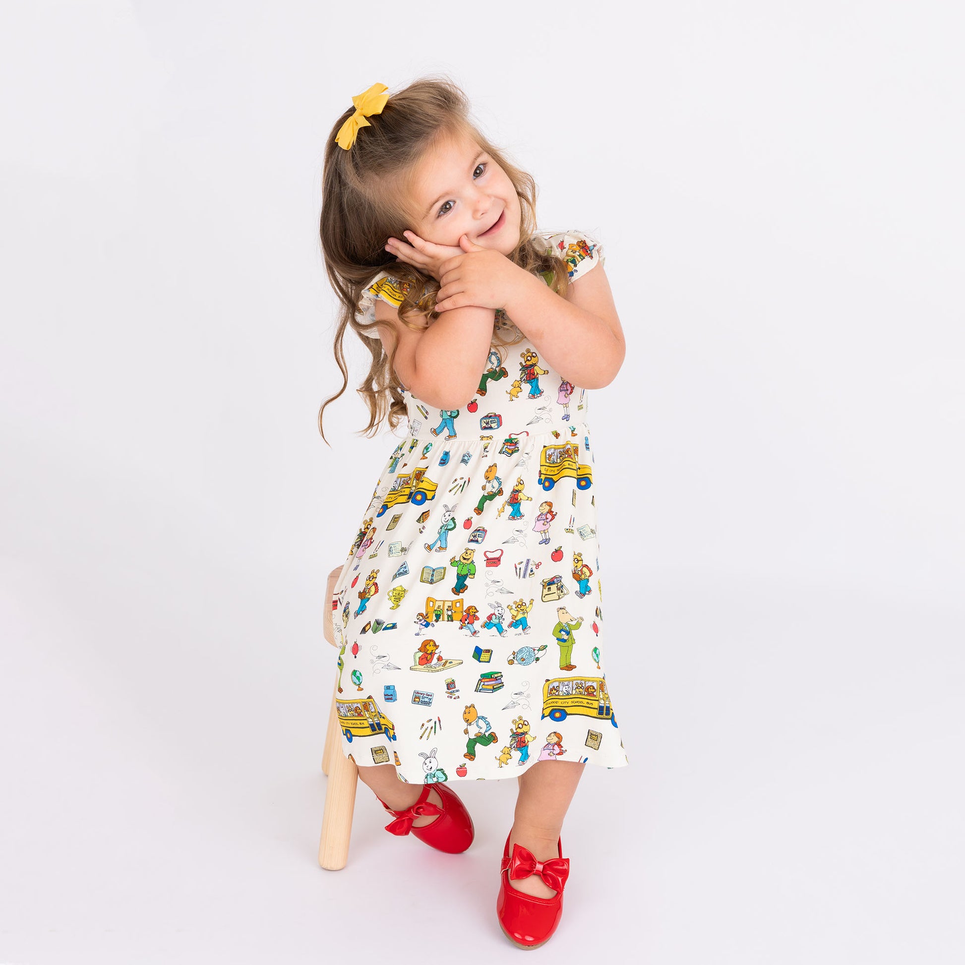 Toddler girl sitting in a chair wearing our twirl bamboo dress featuring playful illustrations of Arthur™ PBS Character, books, and school bus, a beige background.