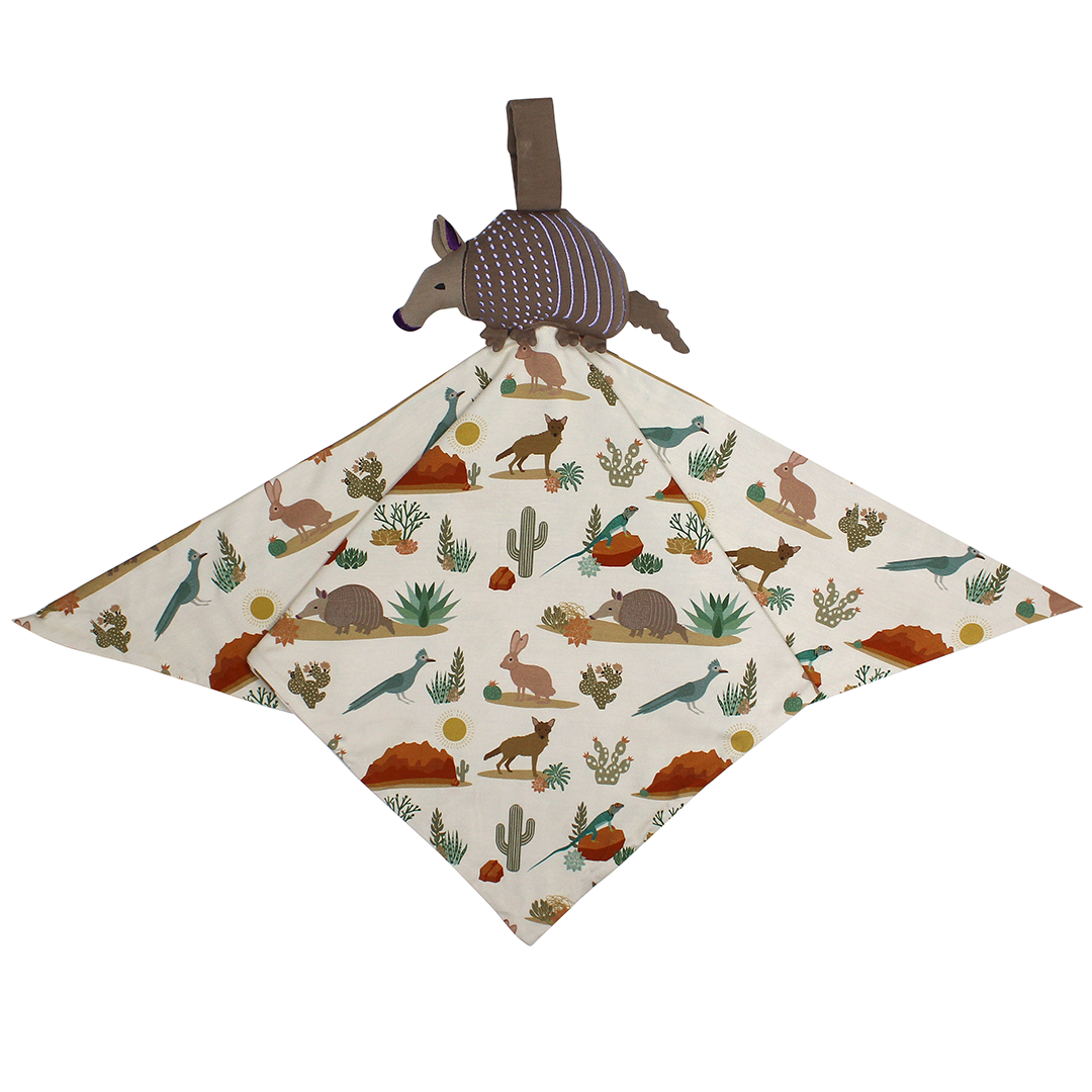 the "desert friends" lovey is a "desert friends" blanket with an armadillo attached to the top of it. the "desert friends" print is a scattered pattern of armadillo, cacti, other desert plants, rocks, desert foxes, lizards, and desert birds. this is all on a beige background.